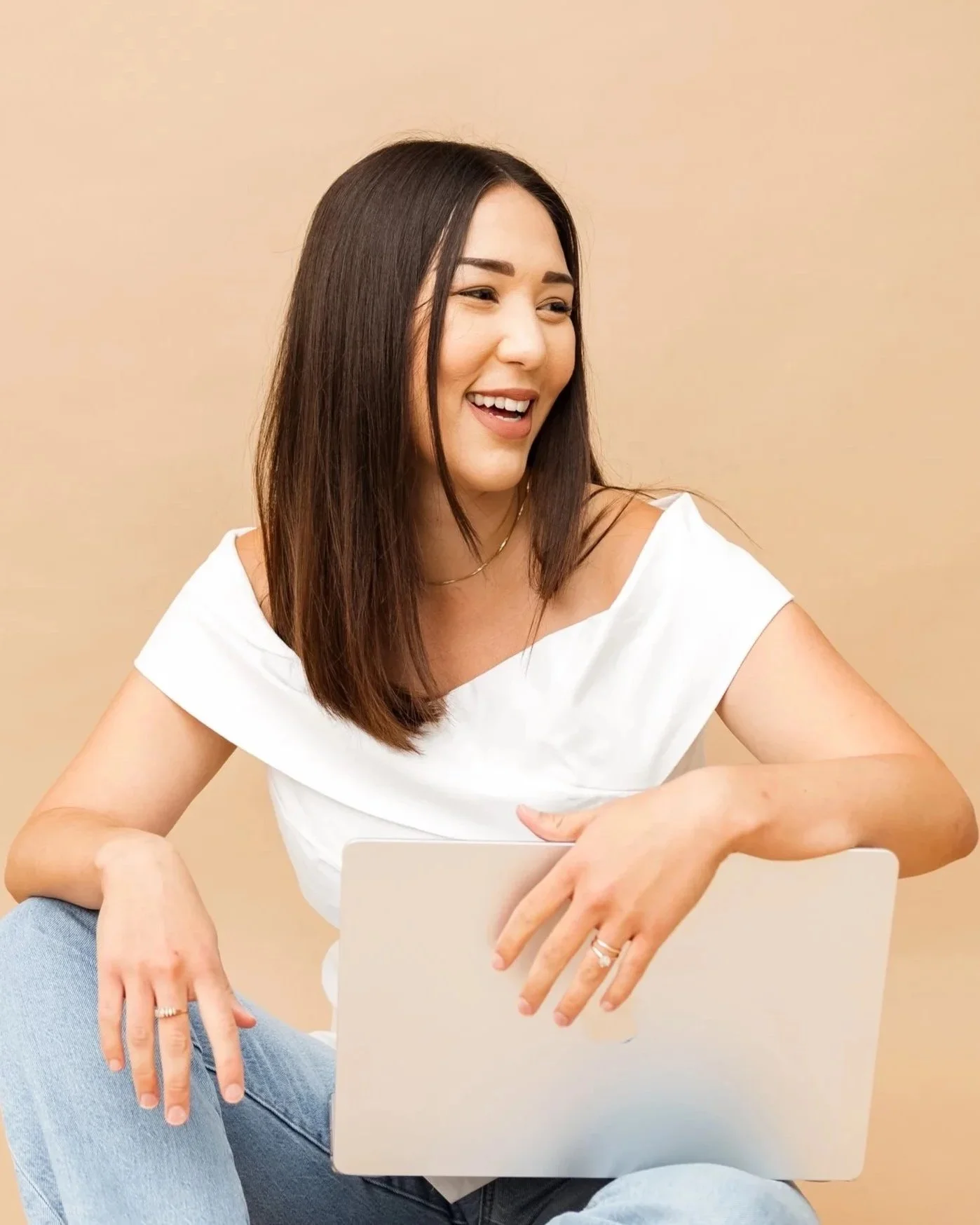 A young woman with dark hair, smiling and looking to her left, sitting cross-legged with a laptop on her lap against a beige background.
