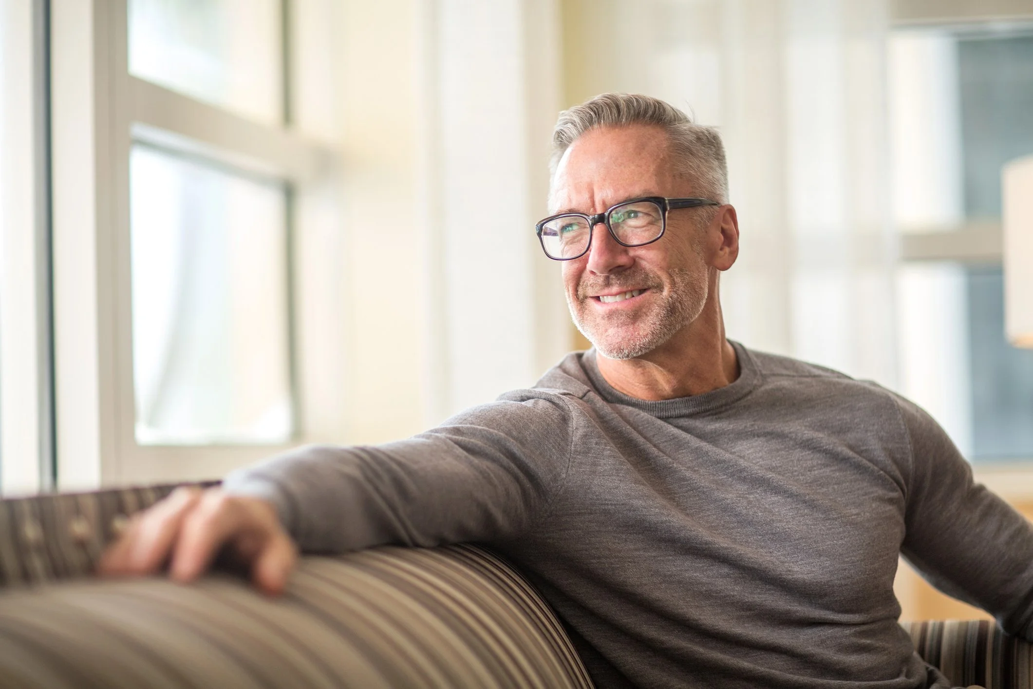 A smiling middle-aged man with gray hair, wearing glasses and a gray long-sleeve shirt, sitting on a striped couch near large windows with sunlight coming through.