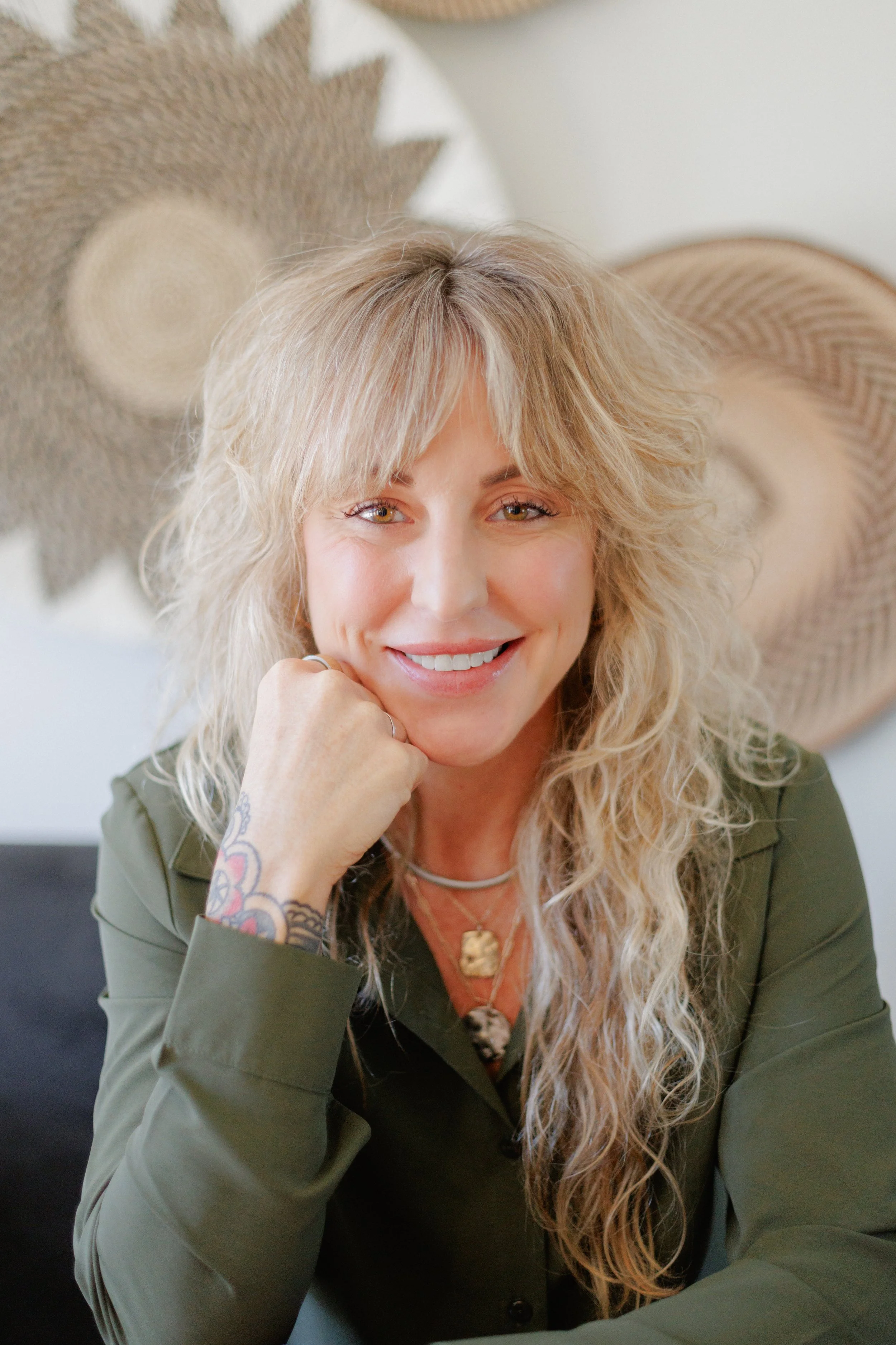 A smiling woman with long curly blond hair, wearing an olive green shirt, sitting indoors with woven wall art in the background.