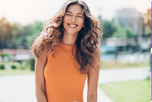A smiling woman wearing a brown scarf and black jacket, holding a to-go coffee cup outdoors in a park with trees and fallen leaves.