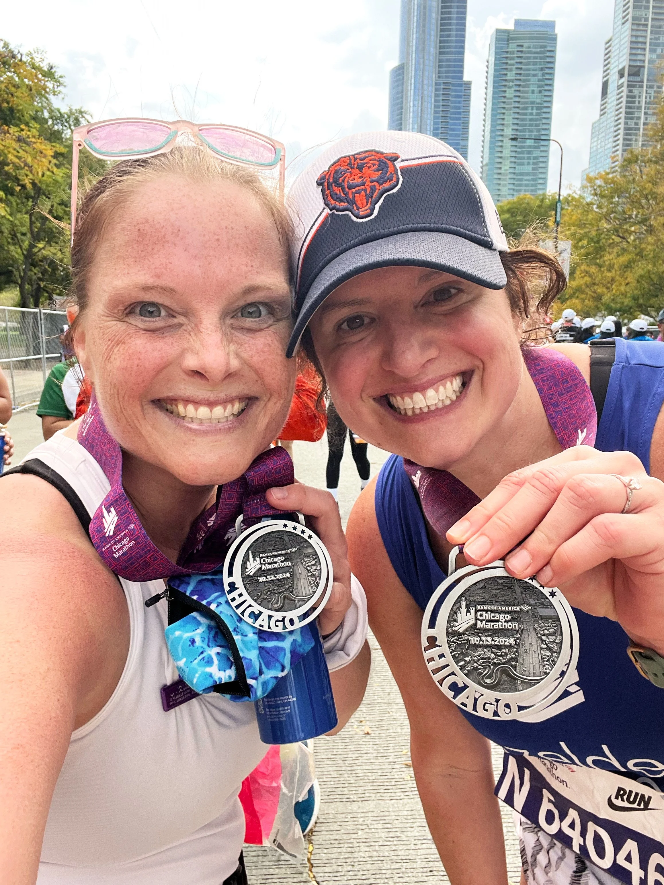 Two female marathon runners smiling and holding their finisher medals at the Chicago Marathon, with city skyscrapers and other runners in the background.
