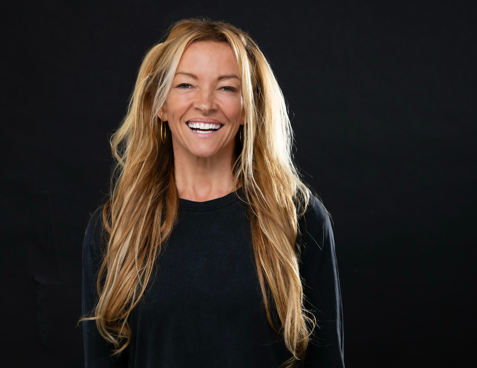 A woman with long blond hair smiling, wearing a black top, against a dark background.