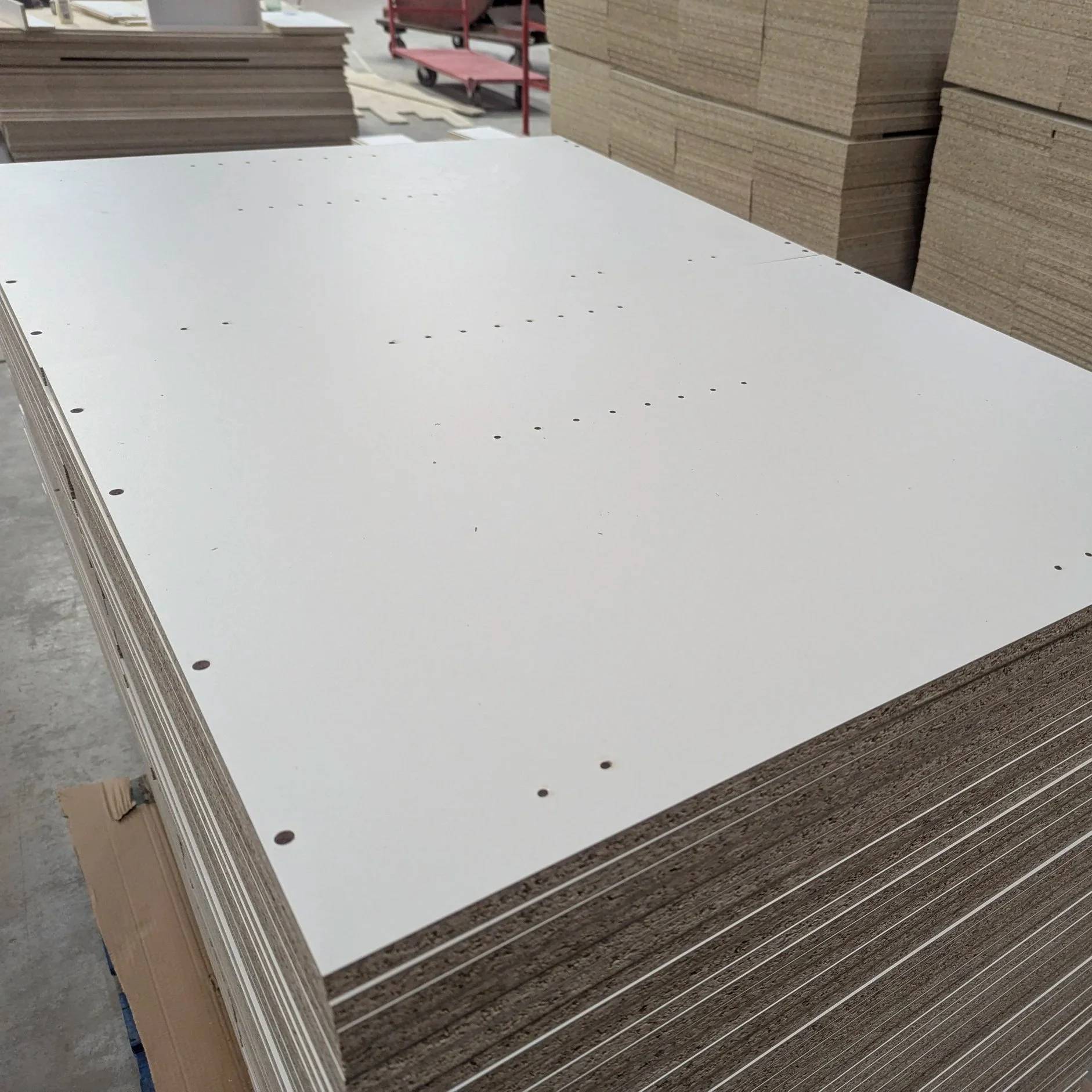 Stack of white drywall sheets with pre-drilled holes in a warehouse or construction site.
