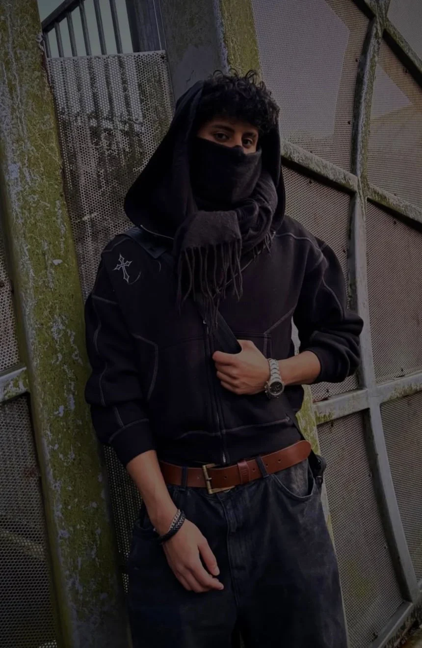 A person with curly hair wearing a black face mask, black hoodie, black jacket with a star emblem on the sleeve, black jeans, brown belt, and a watch, standing against a moss-covered wall and industrial metal gates.