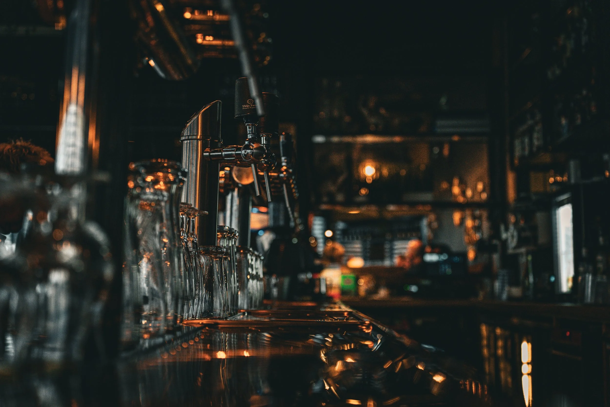 Close-up van een zwart geverfde bar met alcoholglazen en tapkranen onder warme, sfeervolle verlichting.