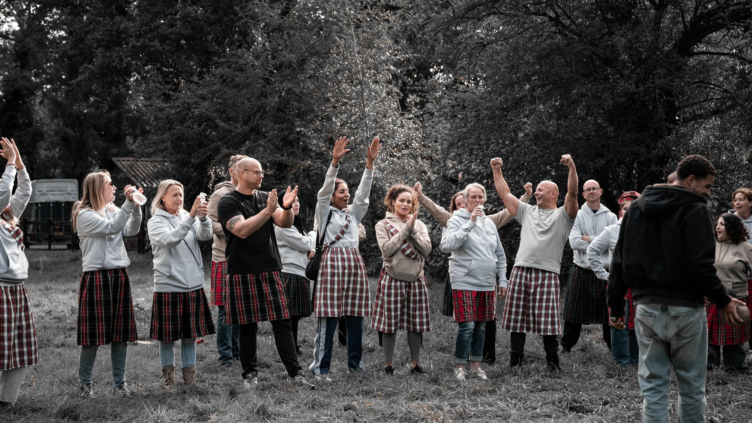 Mensen die in een veld dansen en vieren, gekleed in traditionele Schotse rokken en casual kleding, met een bos op de achtergrond.