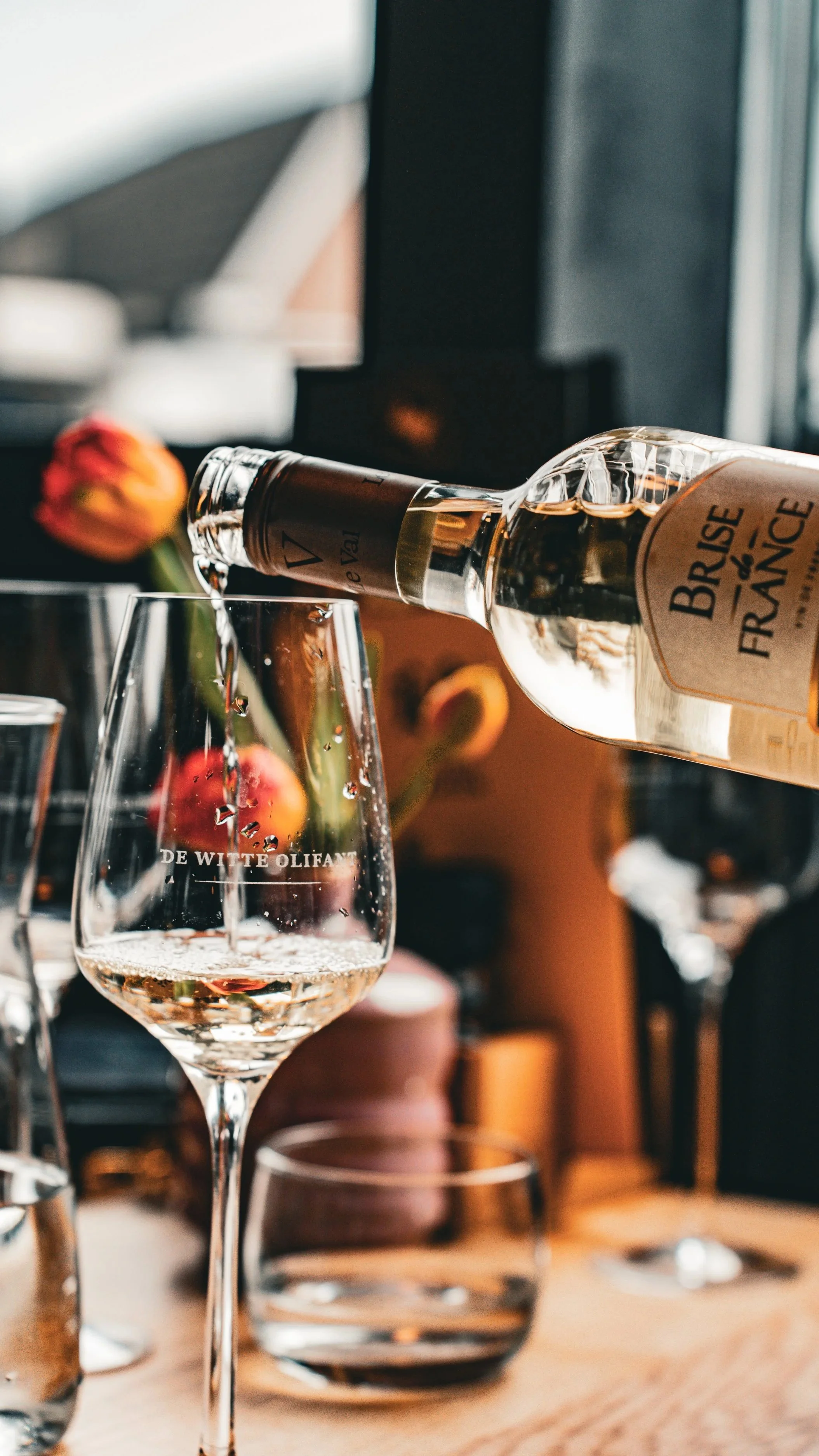 Een glas witte wijn wordt gevuld vanuit een fles, er liggen andere glazen en een vaas met bloemen op de achtergrond.