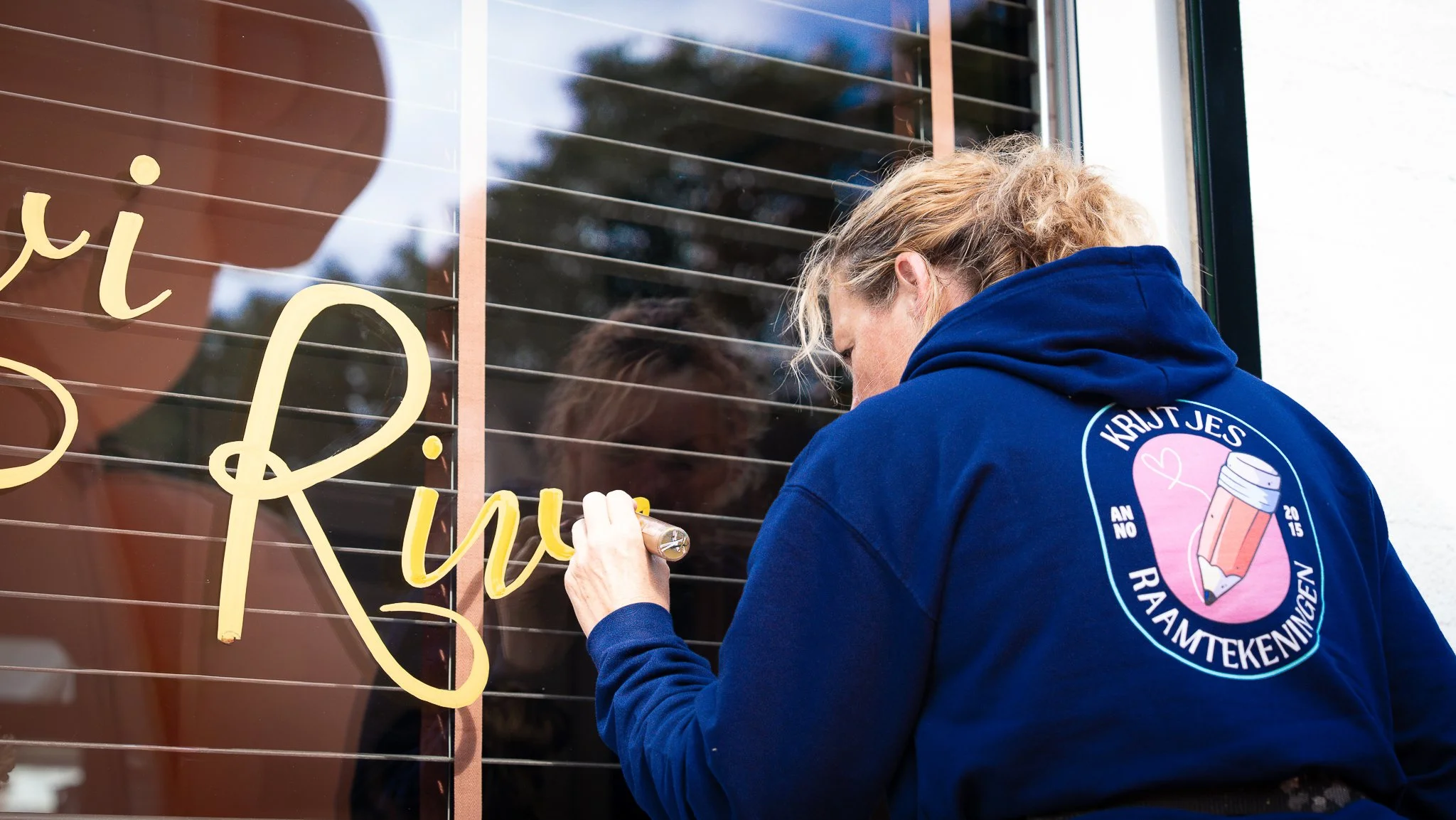Vrouw schrijft met goudkleurige verf op een raam met horizontale lamellen, terwijl ze binnenin naar haar kijkt.