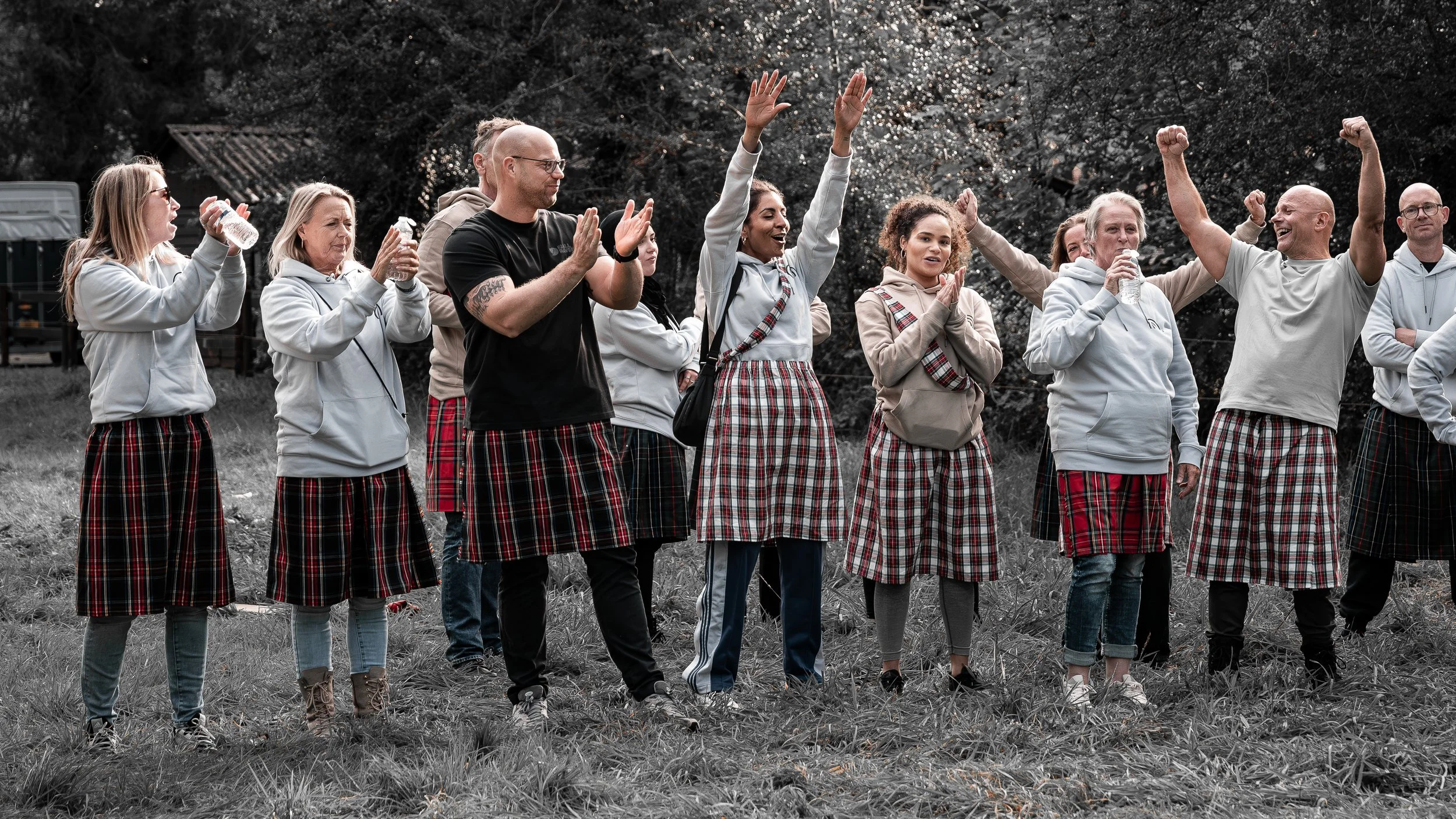 Mensen die in een veld staan, gekleed in traditionele Schotse tartan-kostuums, vieren feest, sommige mensen dansen, anderen drinken water, op de achtergrond bomen en een houten structuur.
