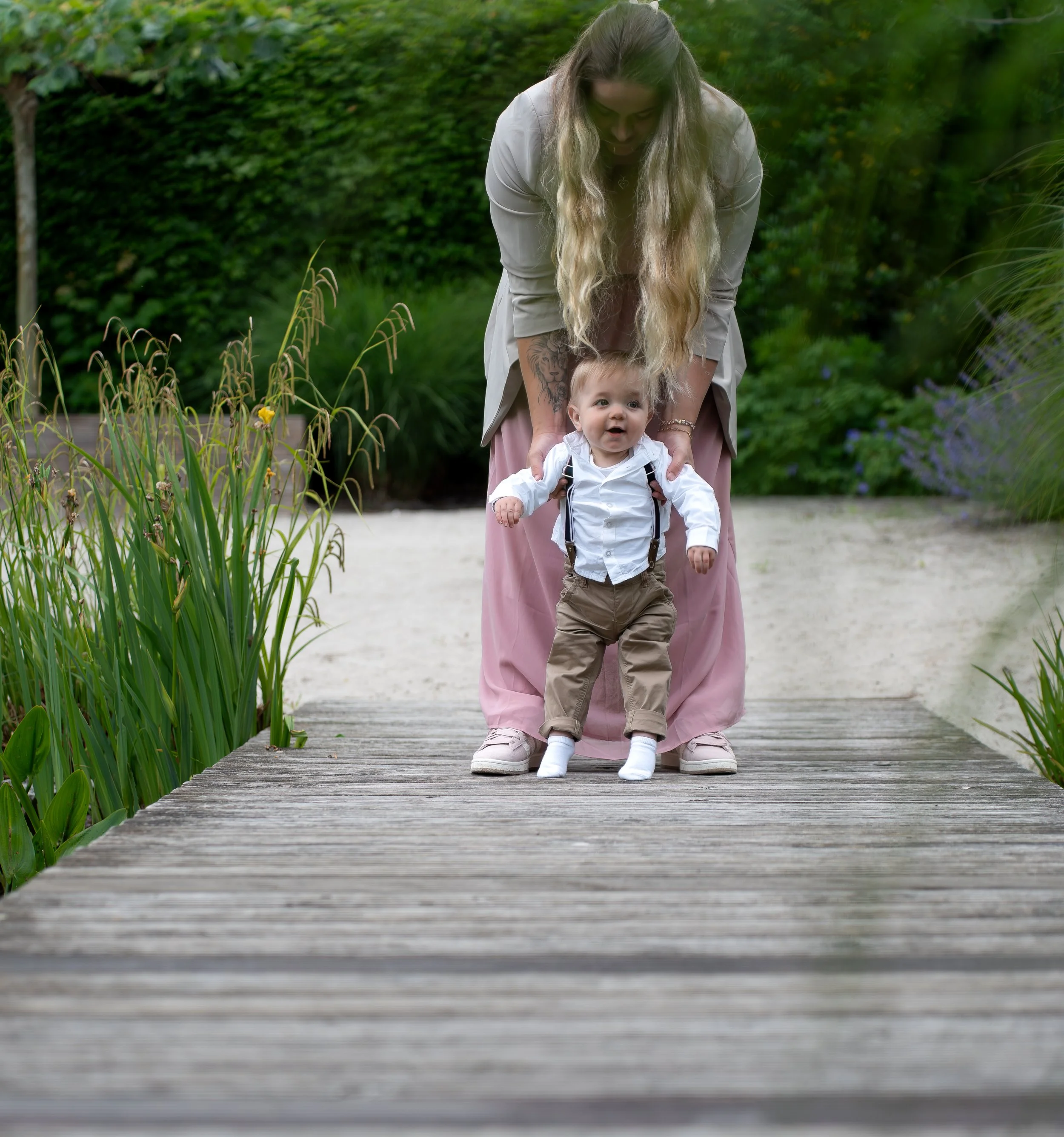 Een jonge vrouw helpt een baby om te lopen op een houten pad in de natuur, met groen en bloemen op de achtergrond.