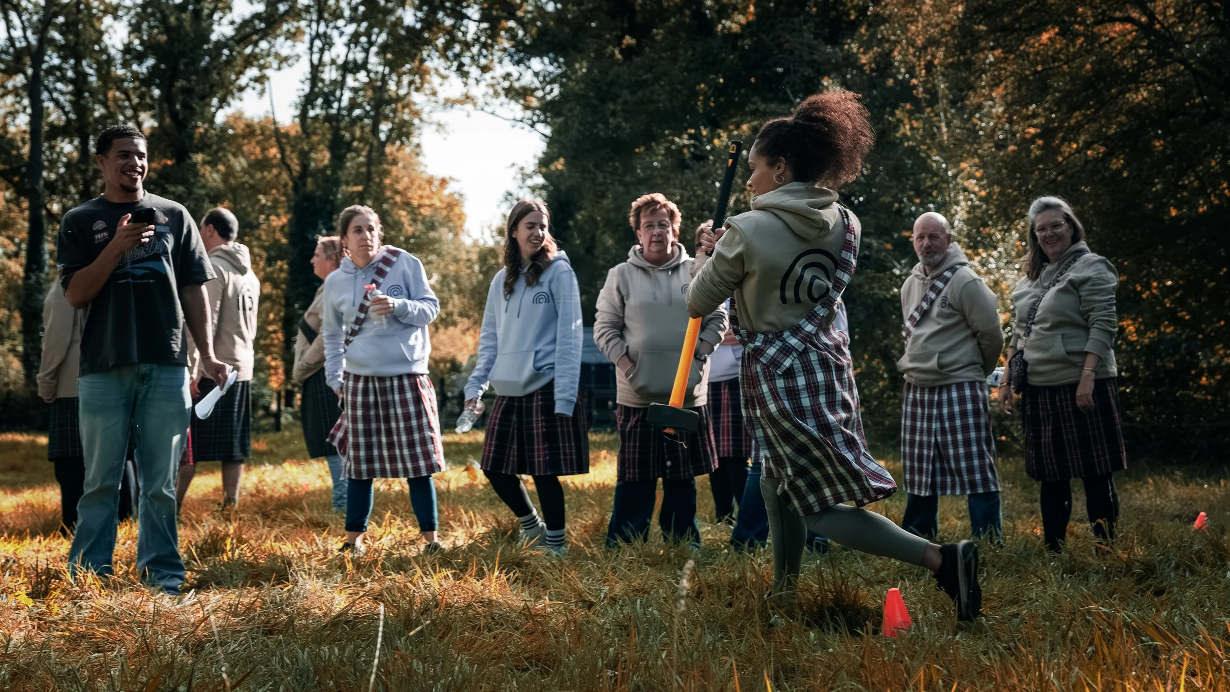 Een groep mensen staat in een bos tijdens de herfst. Ze dragen all-weather kleding met ruitenpatronen en kijken naar een vrouw die met een bijl of hamer staat te wachten, mogelijk voor een activiteit of demonstratie.