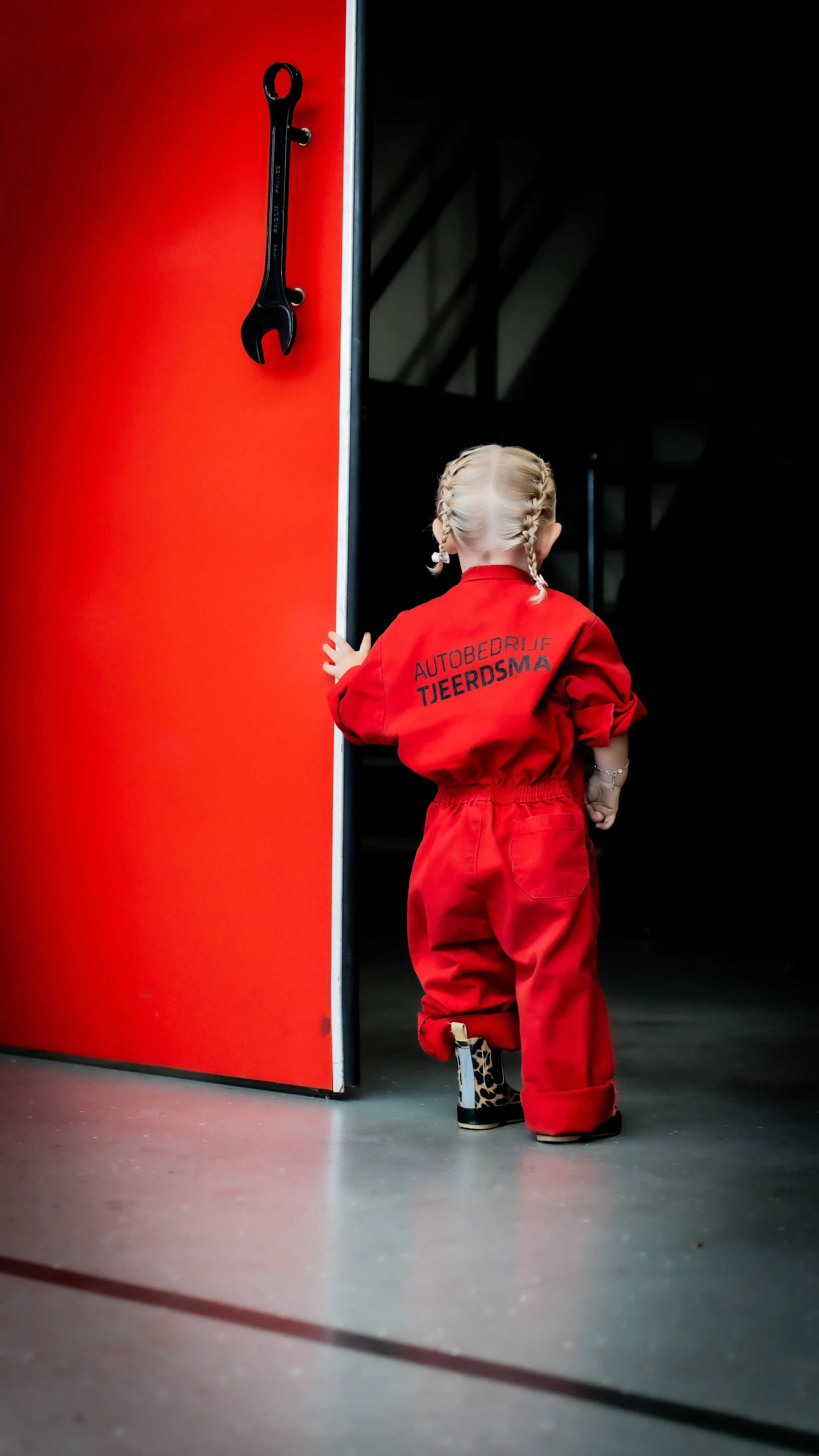 Een jonge meisje in een rood brandweerkostuum staat bij een grote rode deur die een open deur is. Ze kijkt naar binnen in wat waarschijnlijk een garage of werkplaats.