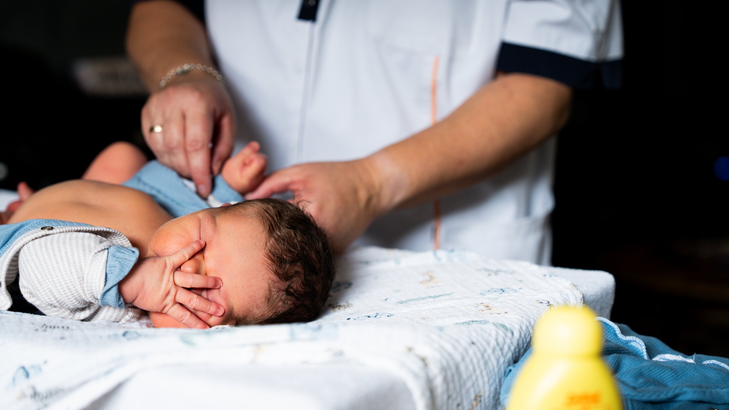 Een pasgeboren baby die op een bed ligt, terwijl een volwassene, mogelijk een arts of een ouder, de baby verzorgt en vasthoudt.