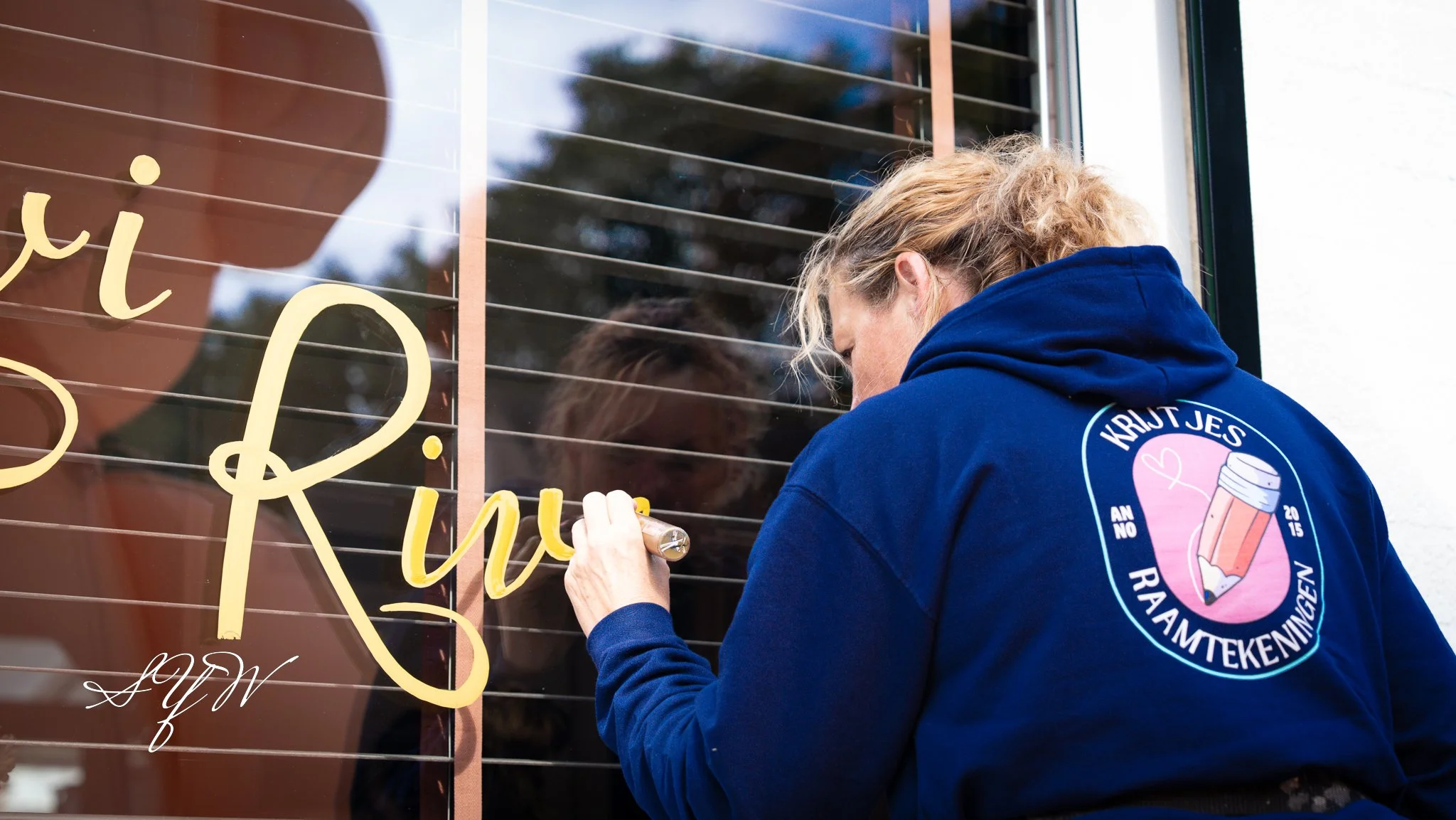 Vrouw draagt een blauwe hoodie met een logo en schrijft met een marker op een raam.