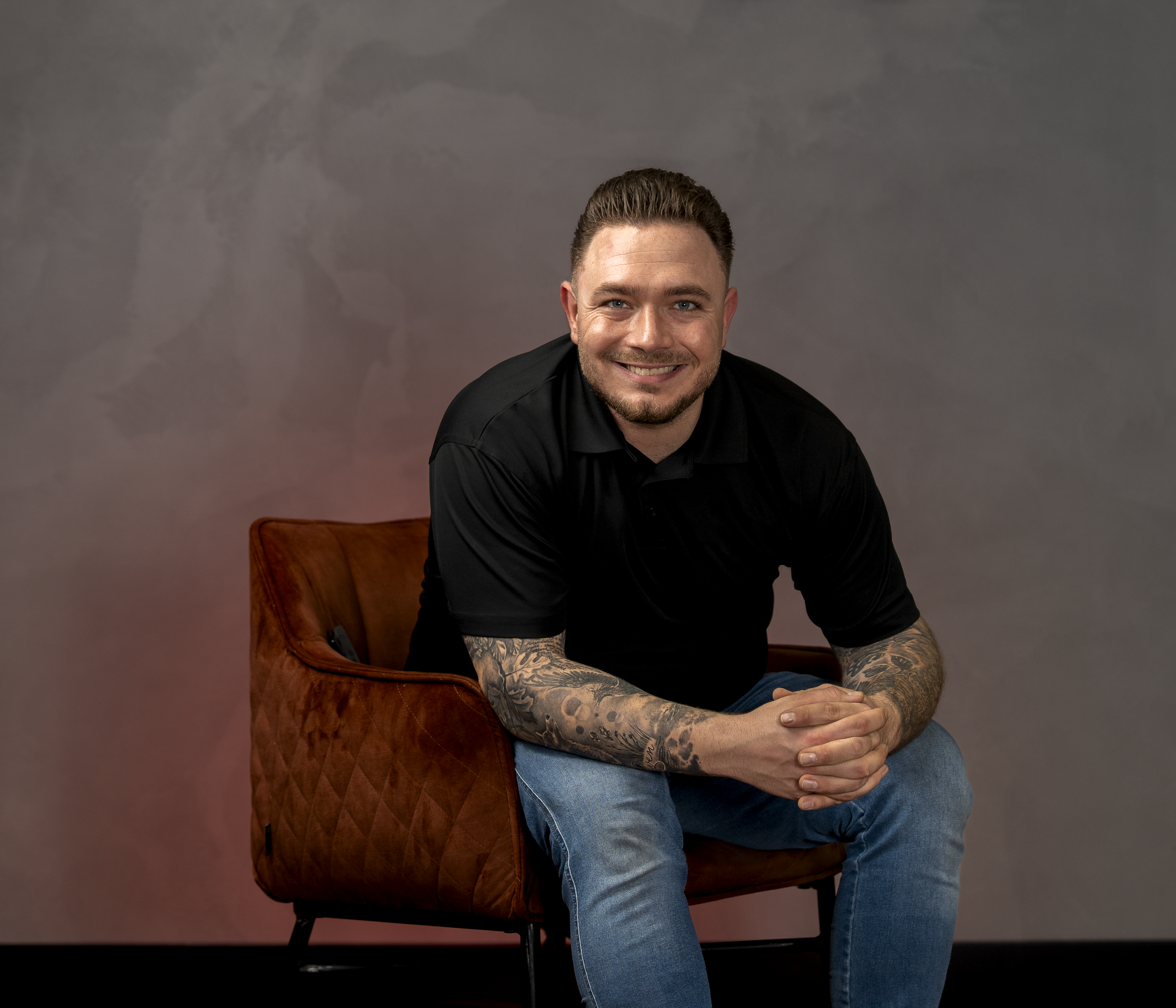 Lachende man met tattoos op armen zittend op een bruine stoel voor een grijze muur.