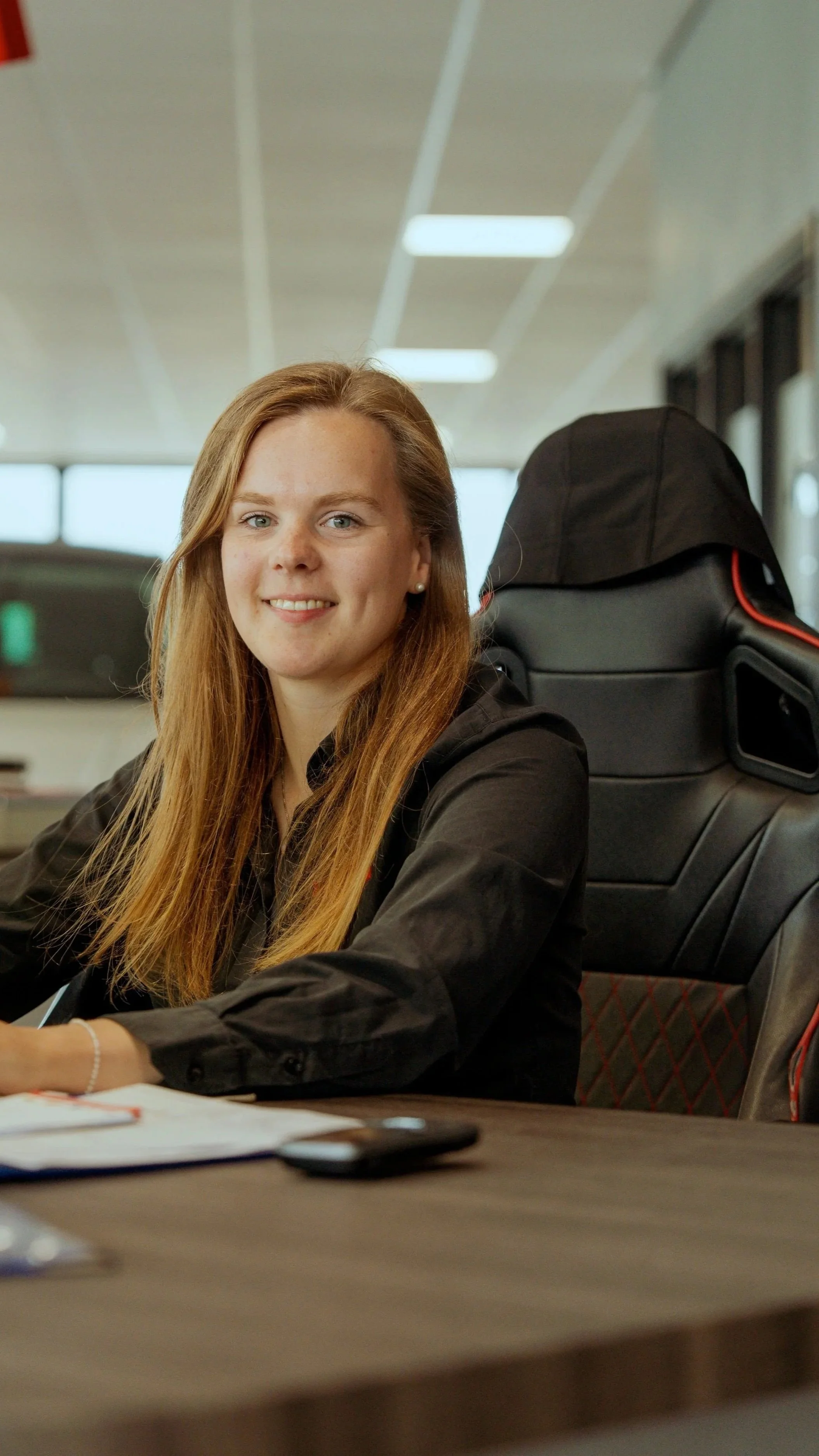 Een glimlachende jonge vrouw met lang rood haar zit aan een tafel in een kantoor, met een gamingstoel op de achtergrond.