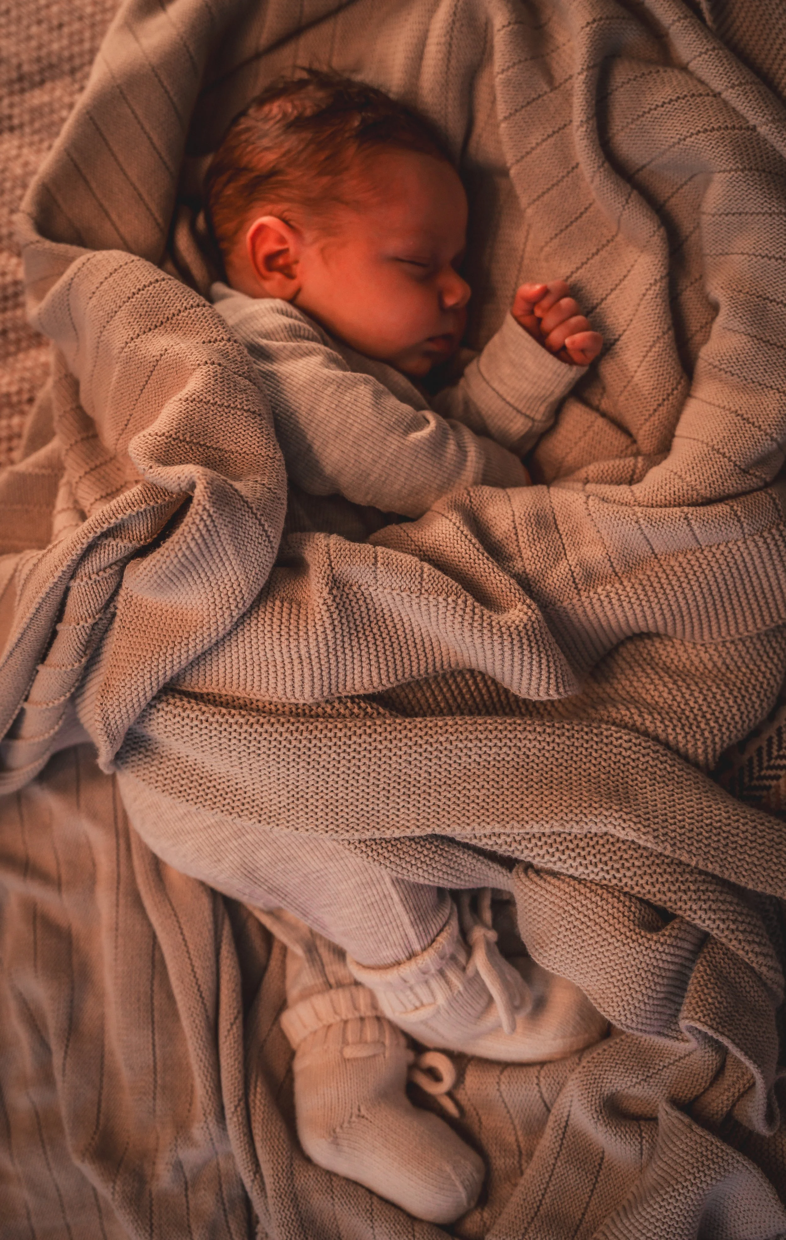 Een slapende baby ingepakt in een beige deken met een ruitpatroon, liggend op een zachte ondergrond.