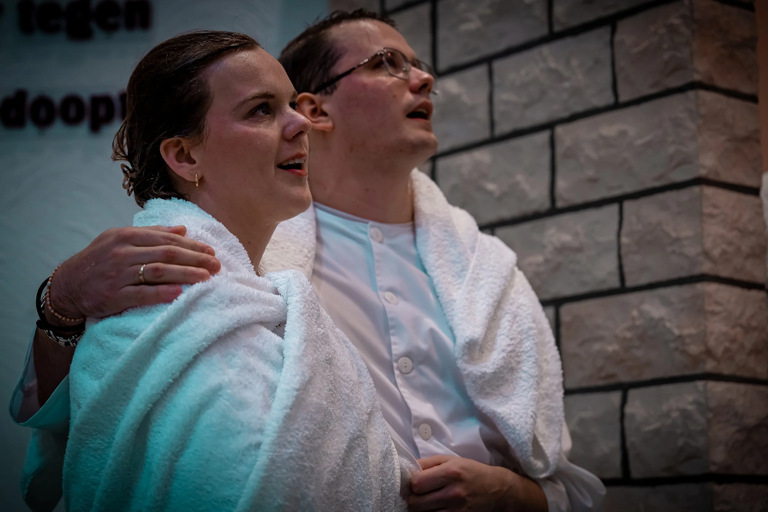 Twee mensen in badjassen, een vrouw en een man, zitten dicht bij elkaar en kijken omhoog met een verraste of verwonderde uitdrukking. Ze zitten in een relaxruimte met een stenen muur op de achtergrond.