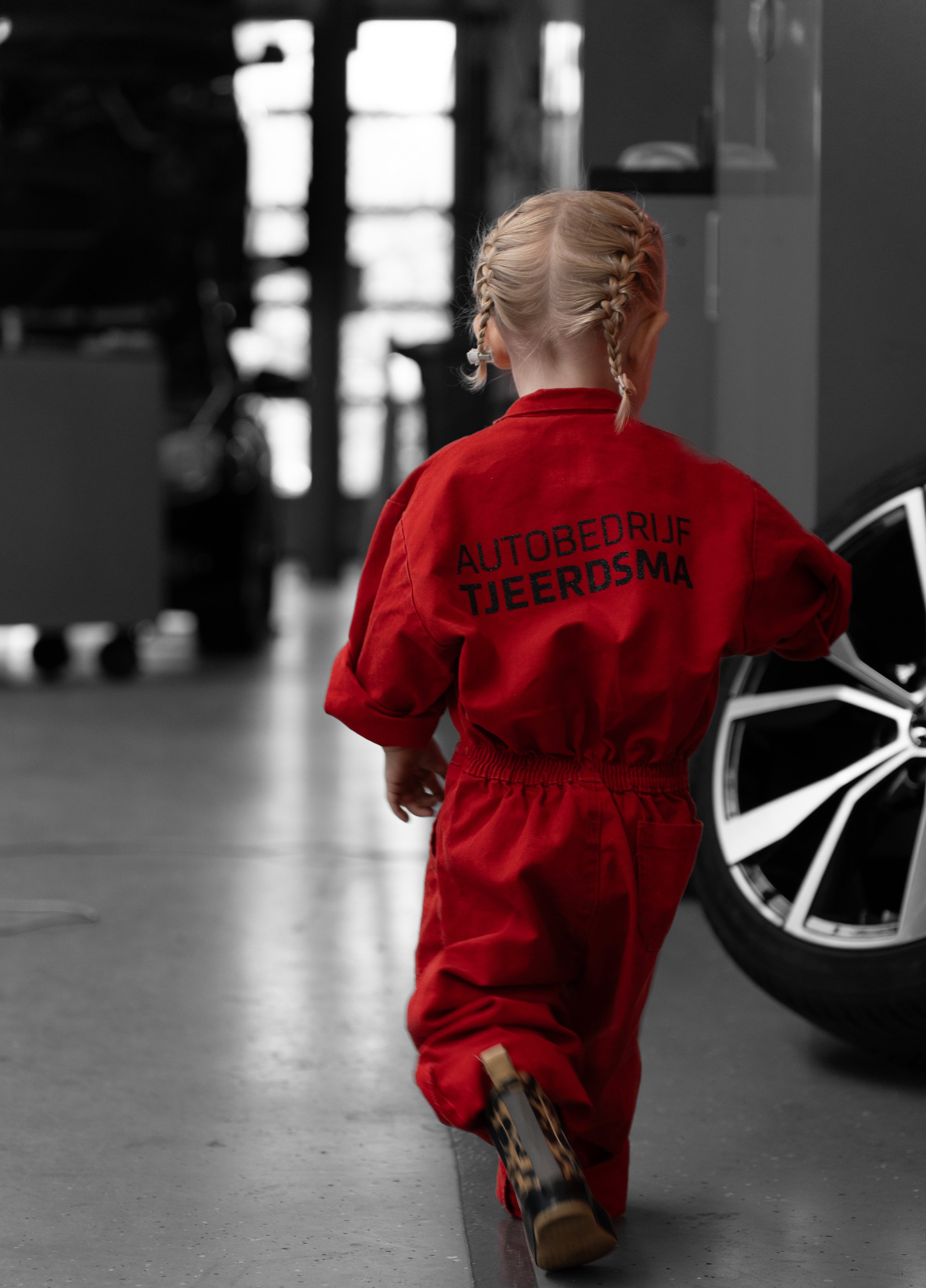 Een jonge vrouw in een rode overalls met de tekst 'Autobedrijf Tjeerdsma' op de achterkant, gezien van achteren in een industriële omgeving, met een auto velg aan de rechterkant.