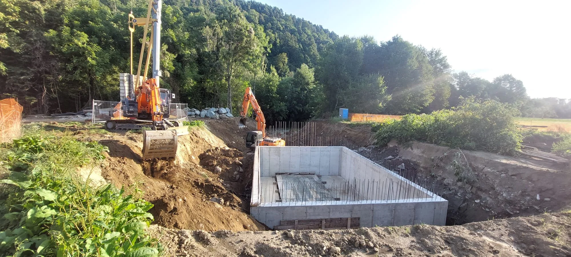 Cantiere edile con due escavatori che lavorano in un sito di costruzione, con una struttura in cemento in fase di realizzazione e un paesaggio naturale circostante.