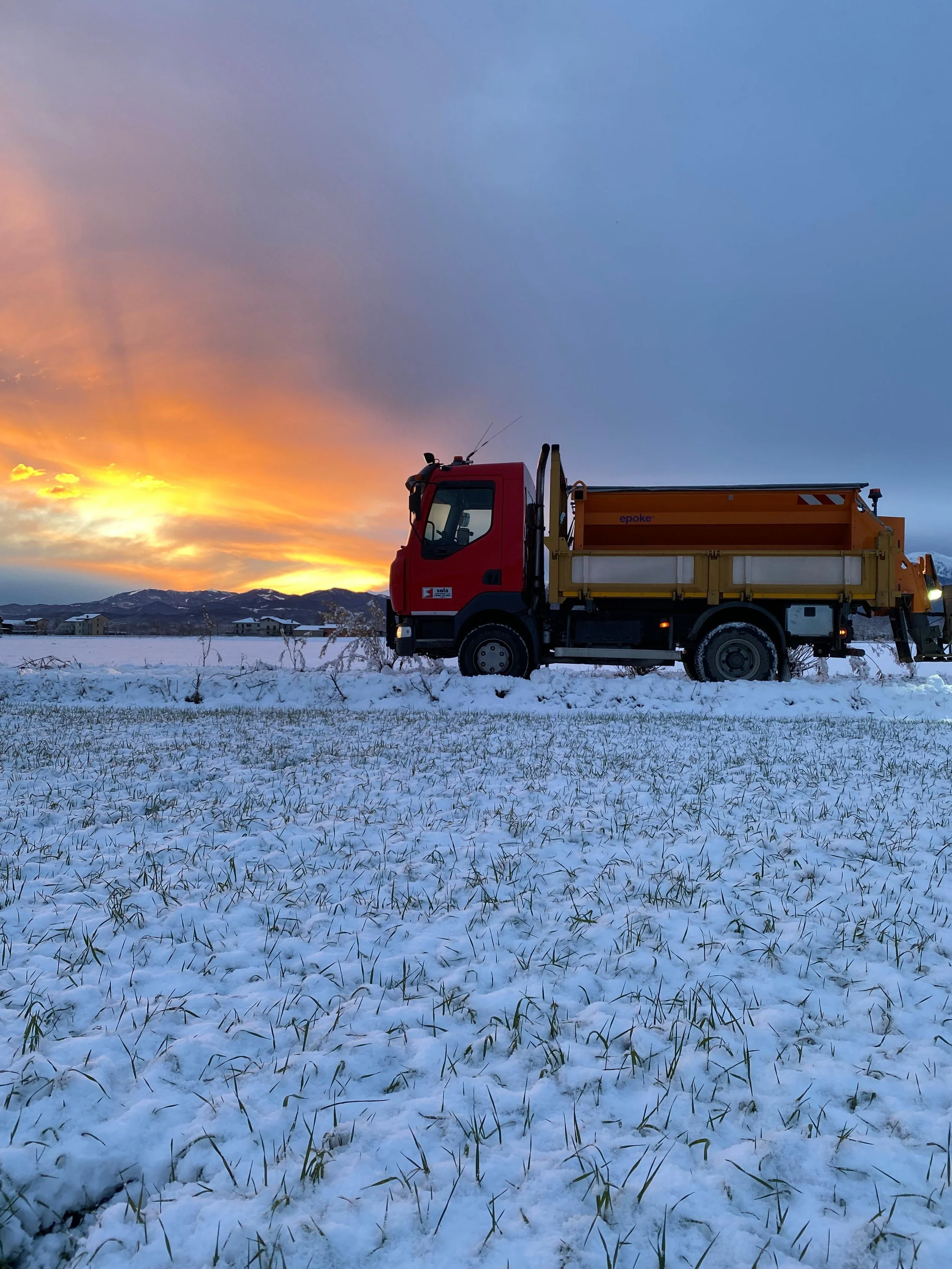 Un camion giallo e rosso parcheggiato su un campo innevato con un tramonto sullo sfondo