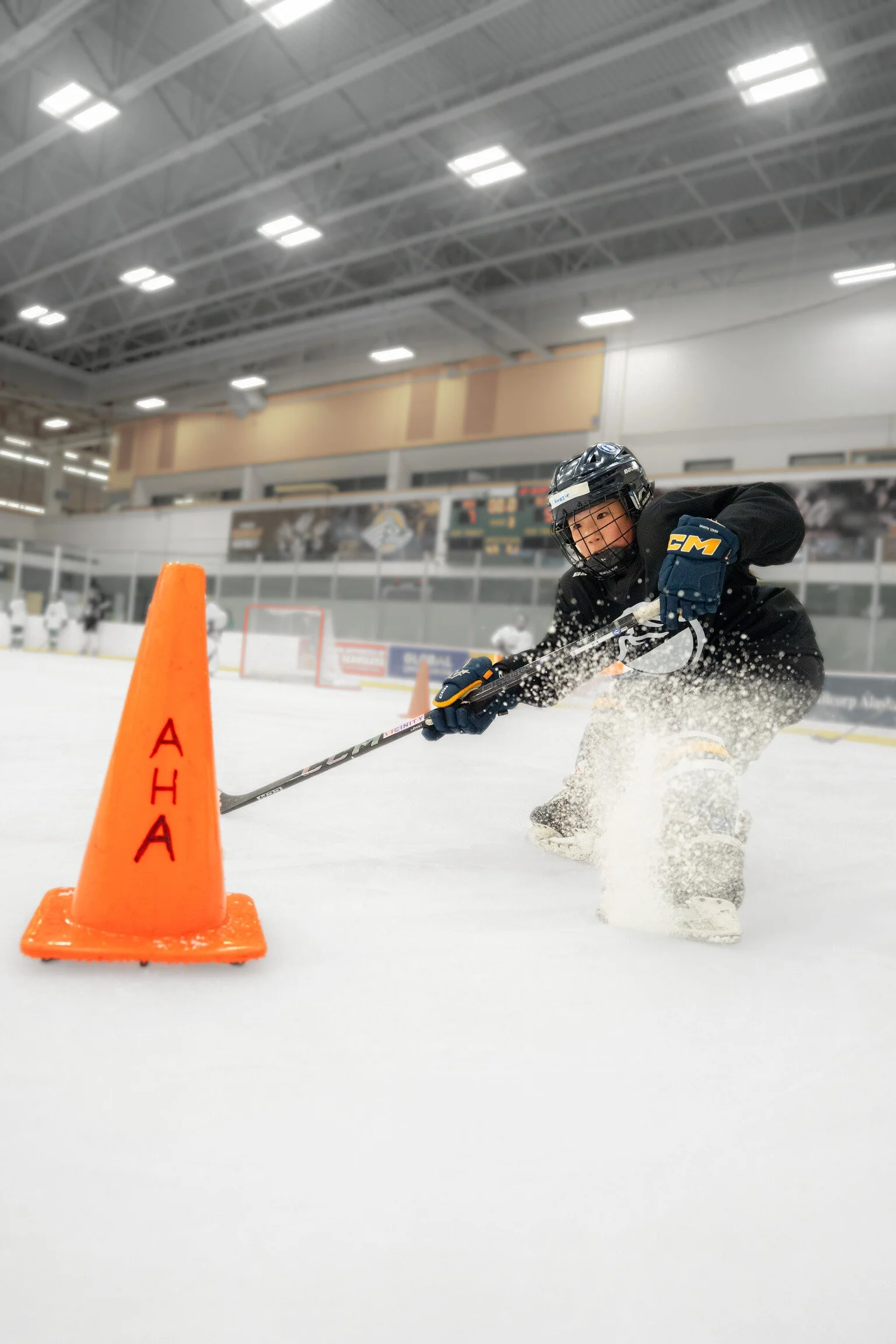 A young hockey player in black uniform and helmet practicing agility drills around an orange cone inside an ice rink.