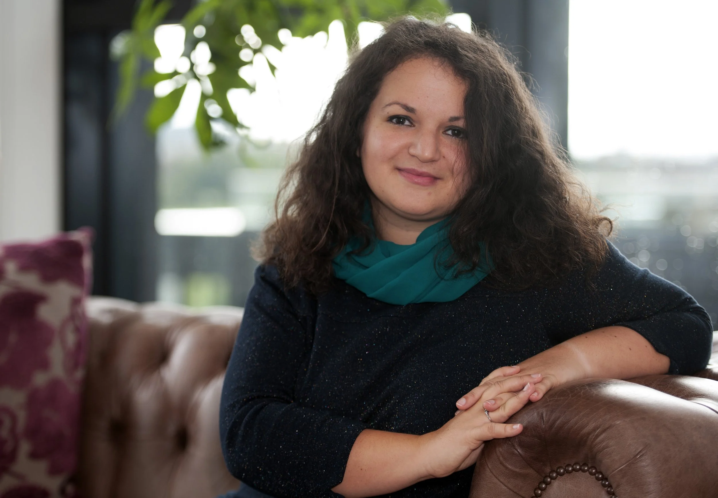 A woman with long curly dark hair, wearing a teal scarf and a dark blue sweater, sitting on a brown leather sofa in a brightly lit room with green plants and large windows in the background.