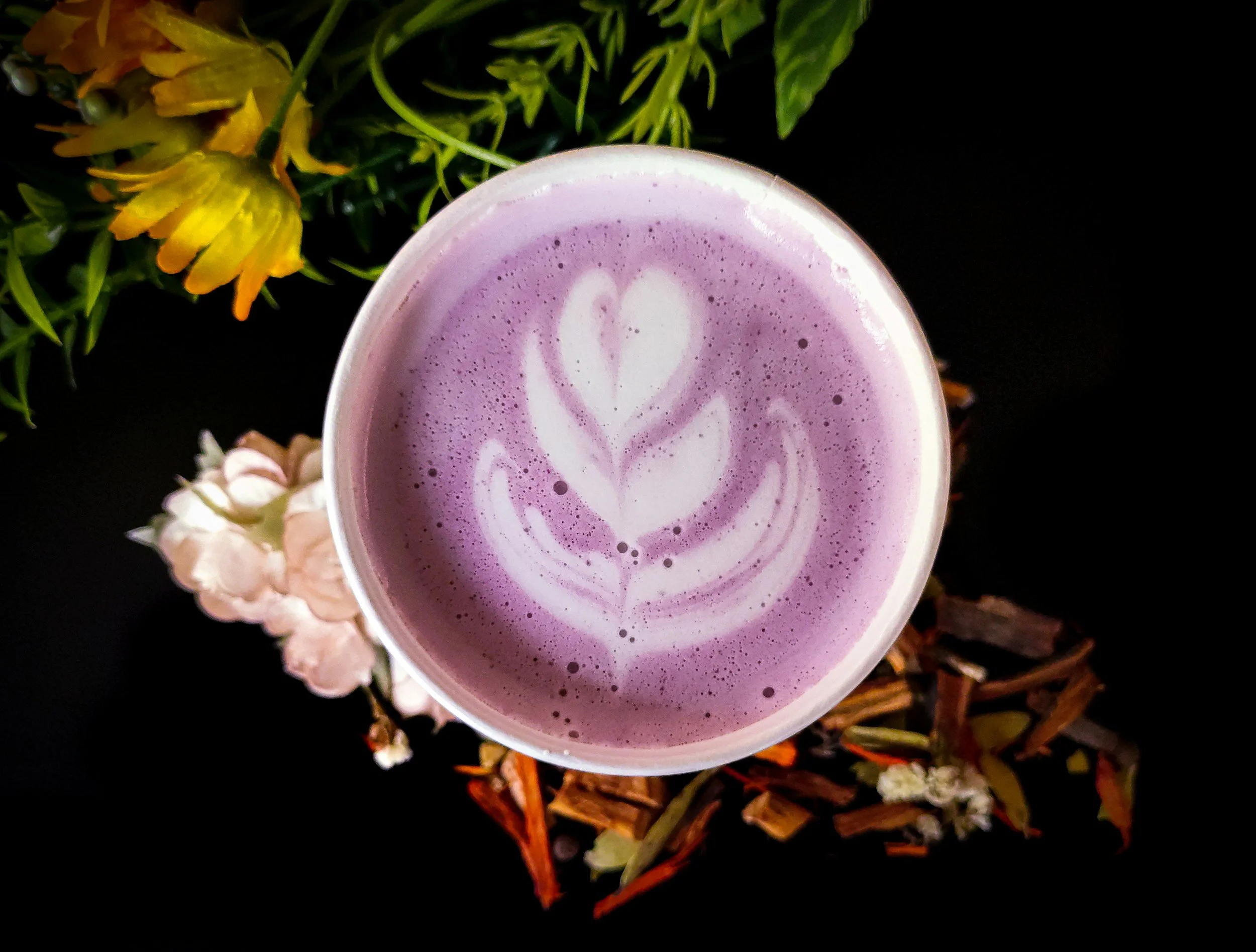 Eine Tasse mit lila Bounty Latte mit Schaumkunst, auf einem schwarzen Hintergrund, umgeben von Blumen und Holzstücken.