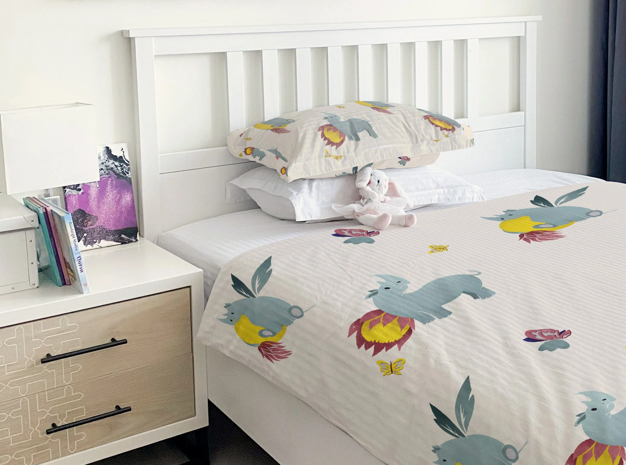 A neatly made bed with a white wooden headboard, featuring unicorn-patterned sheets and pillowcases, a plush bunny toy, and a white nightstand with books and a purple and black abstract art piece.