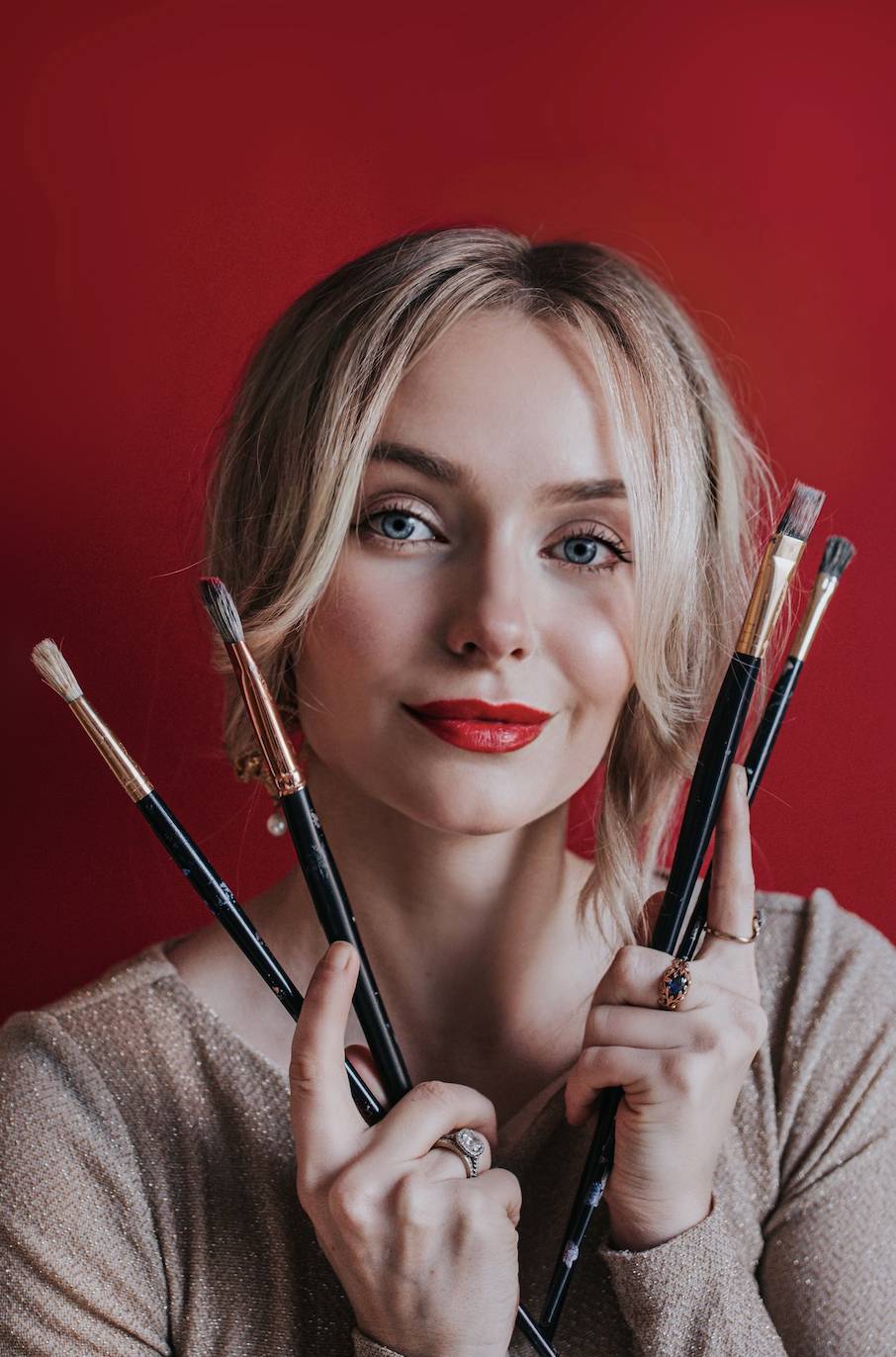 A woman with blonde hair and red lipstick holding several paintbrushes in front of a red background.
