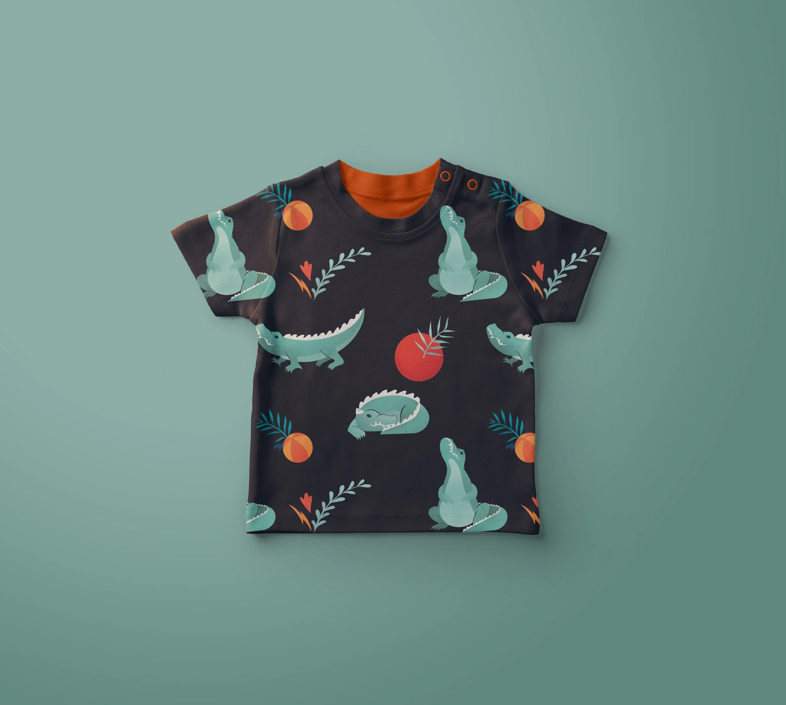Child's dark-colored t-shirt with a pattern of green crocodiles, orange tomatoes, and blue leafy branches, laid flat on a light teal background.