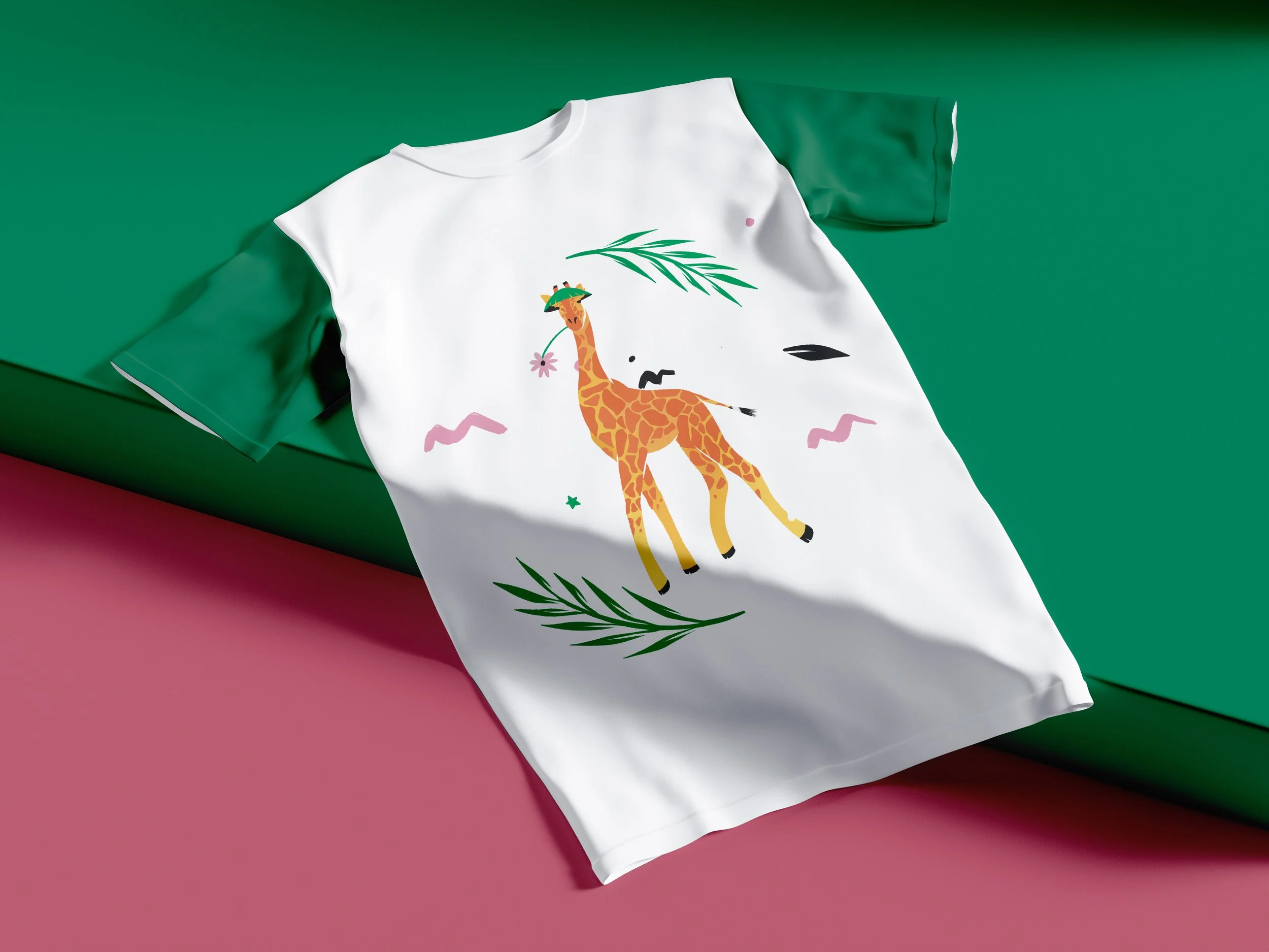White T-shirt with green sleeves featuring a cartoon giraffe wearing a green hat, holding a pink flower in its mouth, surrounded by jungle leaves and abstract shapes, lying on a pink and green background.