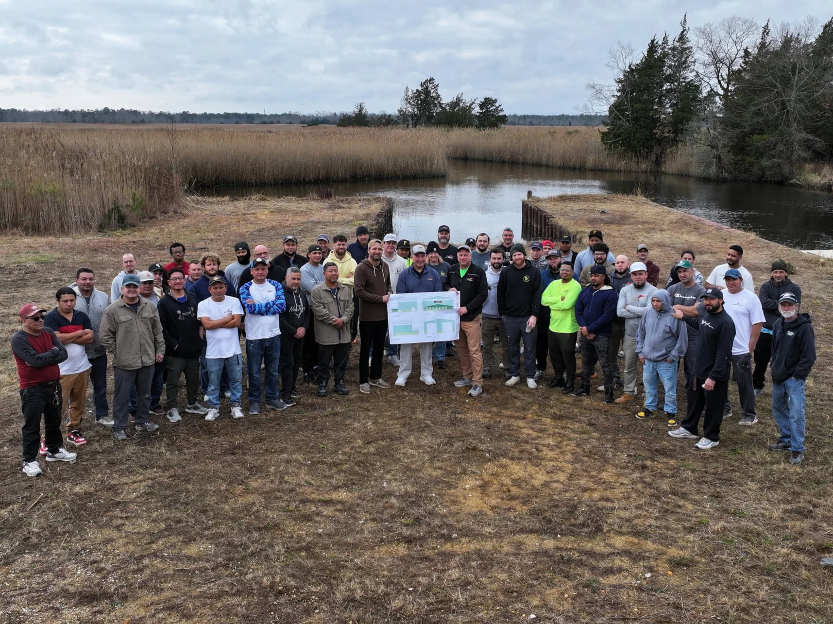 Group of people outdoors near a waterway, holding a large map or blueprint, with trees and reeds in the background on a cloudy day.