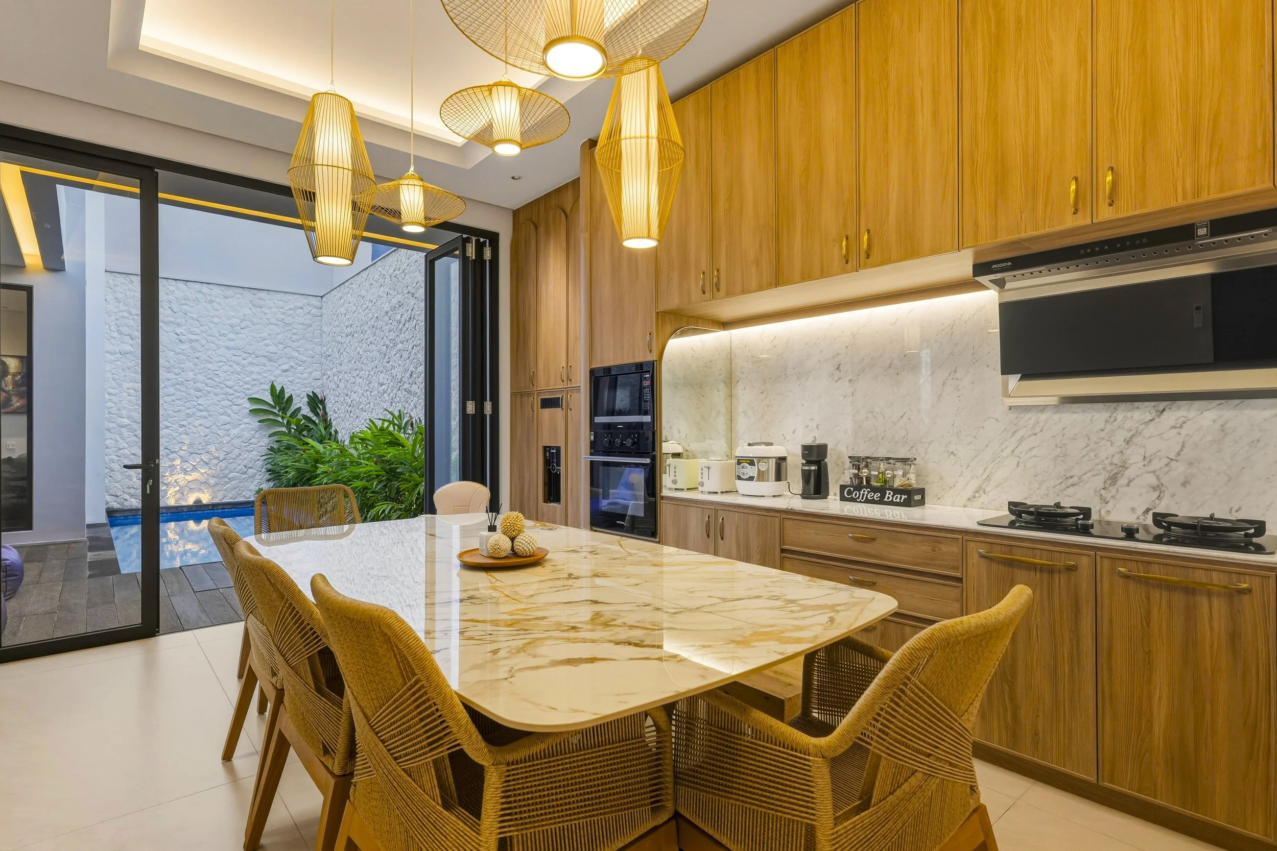 Modern kitchen featuring wooden cabinets, marble countertops, a marble island with wicker chairs, hanging pendant lights, and sliding glass doors opening to an outdoor patio with a white stone wall and green plants.