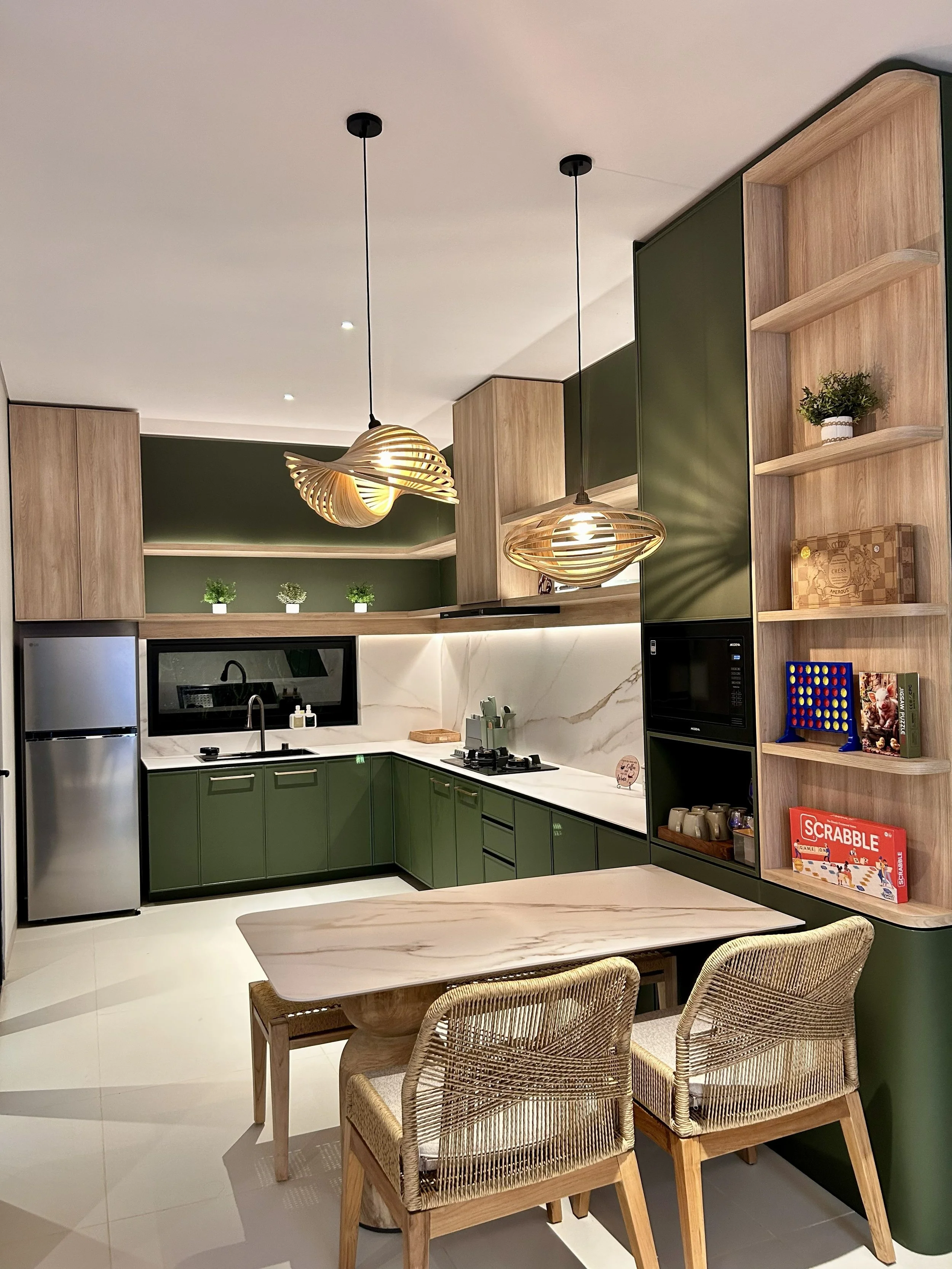 Modern kitchen with green cabinets, wooden open shelves, and artistic pendant lights. There's a small dining table with wicker chairs, and a bookshelf with board games and books.