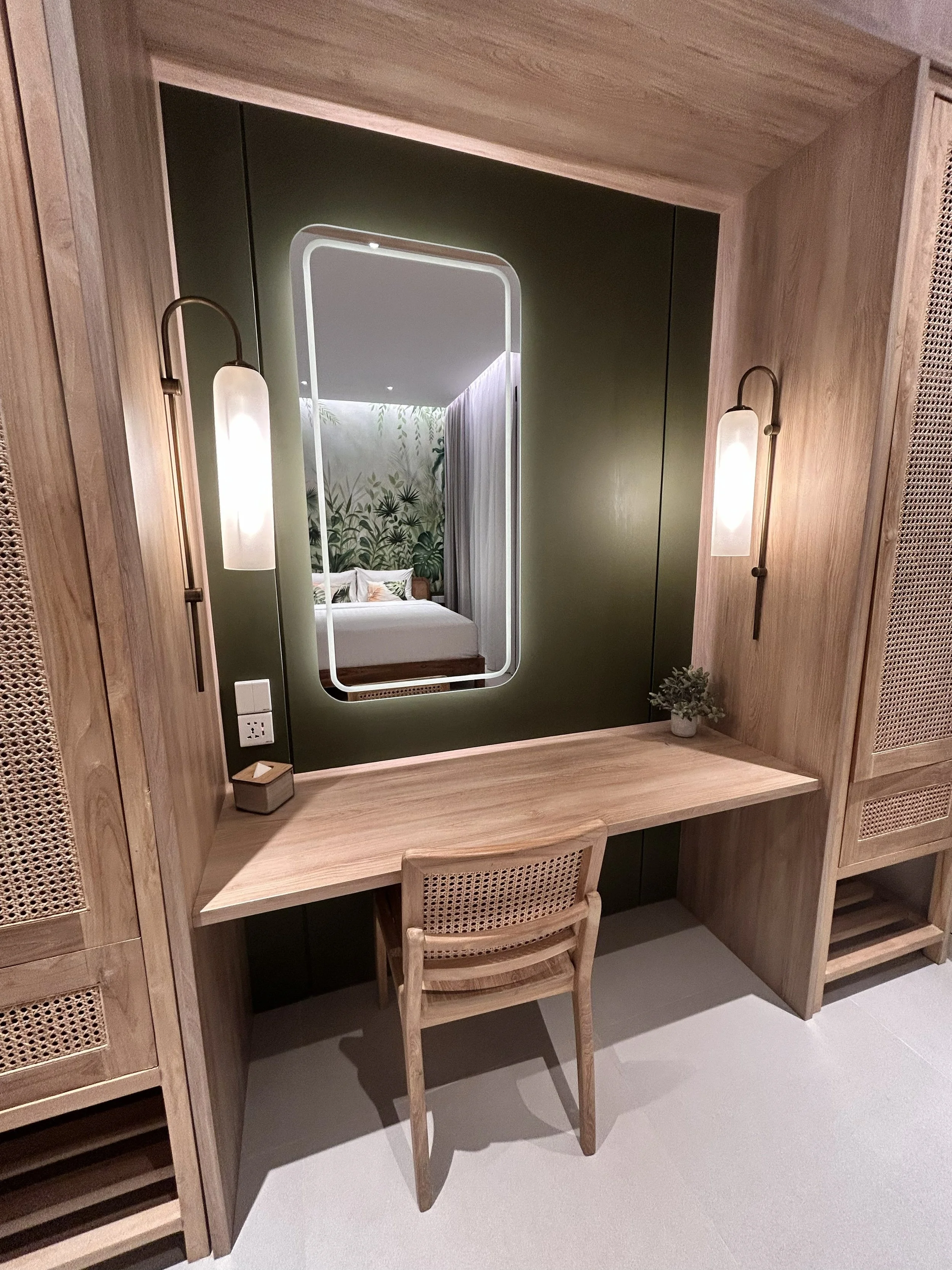 A workspace with a wooden desk and a chair, decorated with a small potted plant and a batik box, and large wall-mounted lamps on either side, with a mirror reflecting a bedroom with a bed, green leafy wallpaper, and curtains.