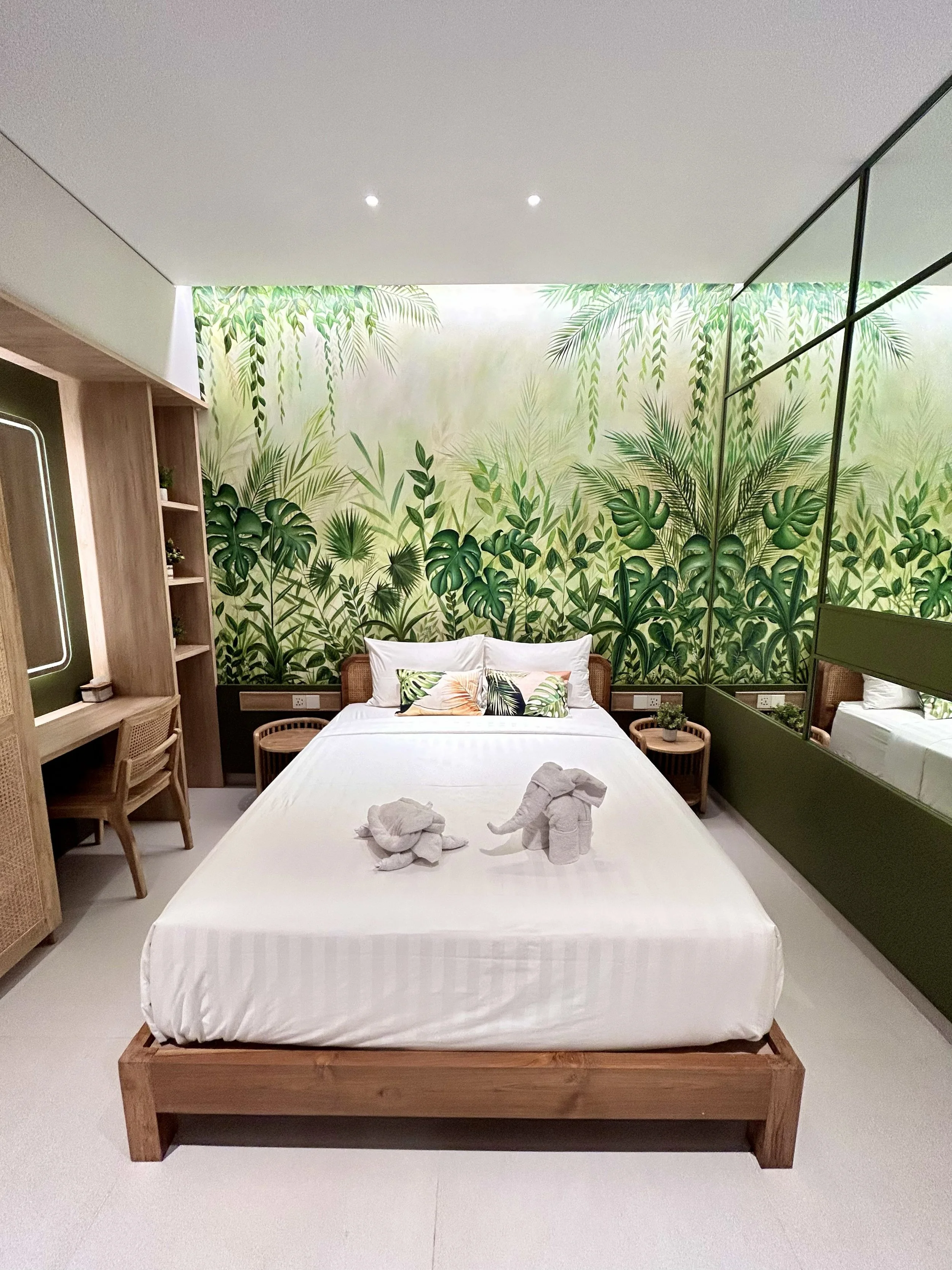 A bedroom with a tropical plant-themed mural wall, a wooden bed frame with white bedding, and decorative elephant towels on the bed. There are small wooden side tables, and a lounging chair next to a built-in wooden shelf with small plants. The room 