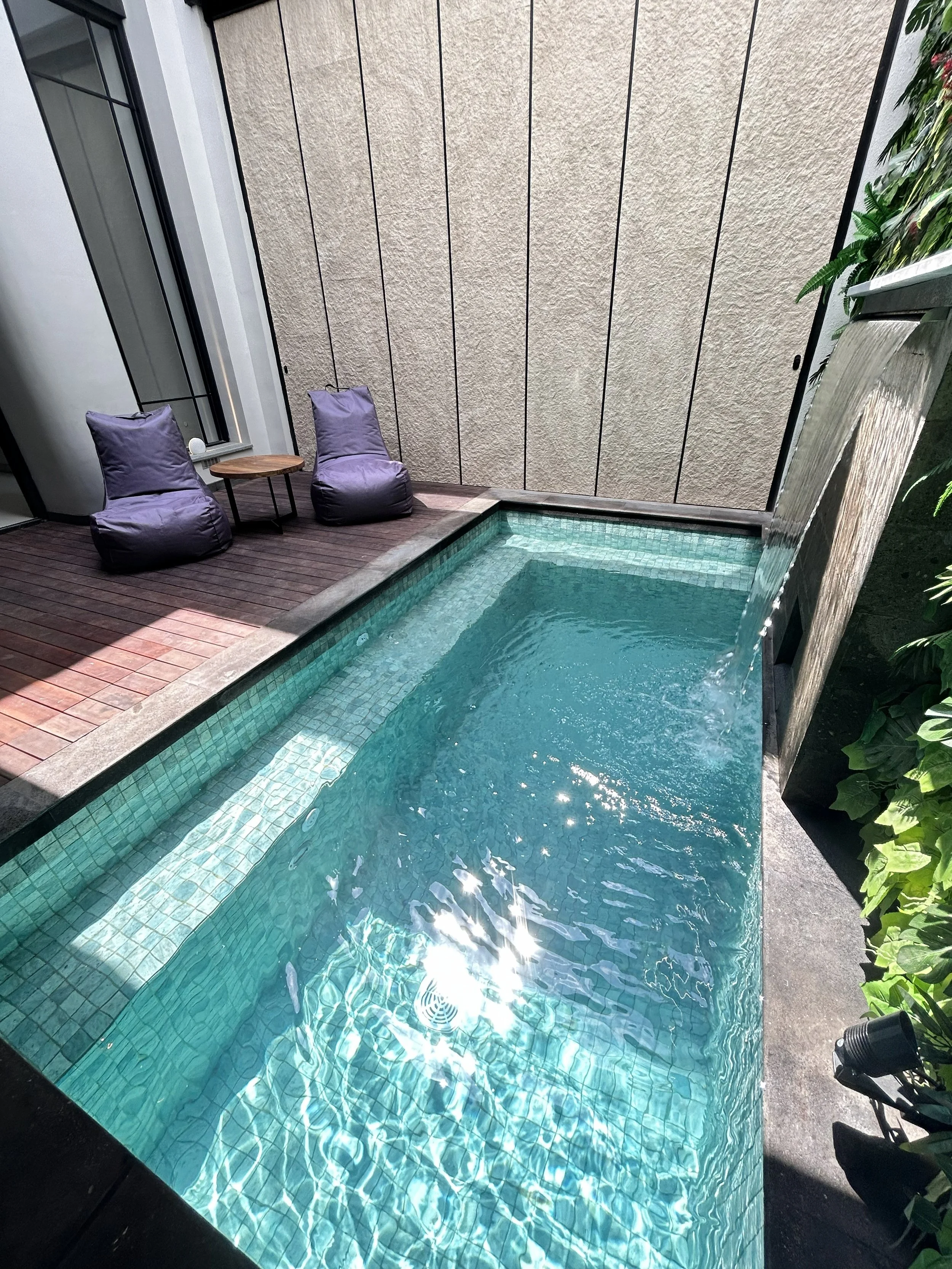A small outdoor pool with clear turquoise water, two purple lounge chairs, a small wooden side table, green plants along the edge, and a textured beige wall in the background.