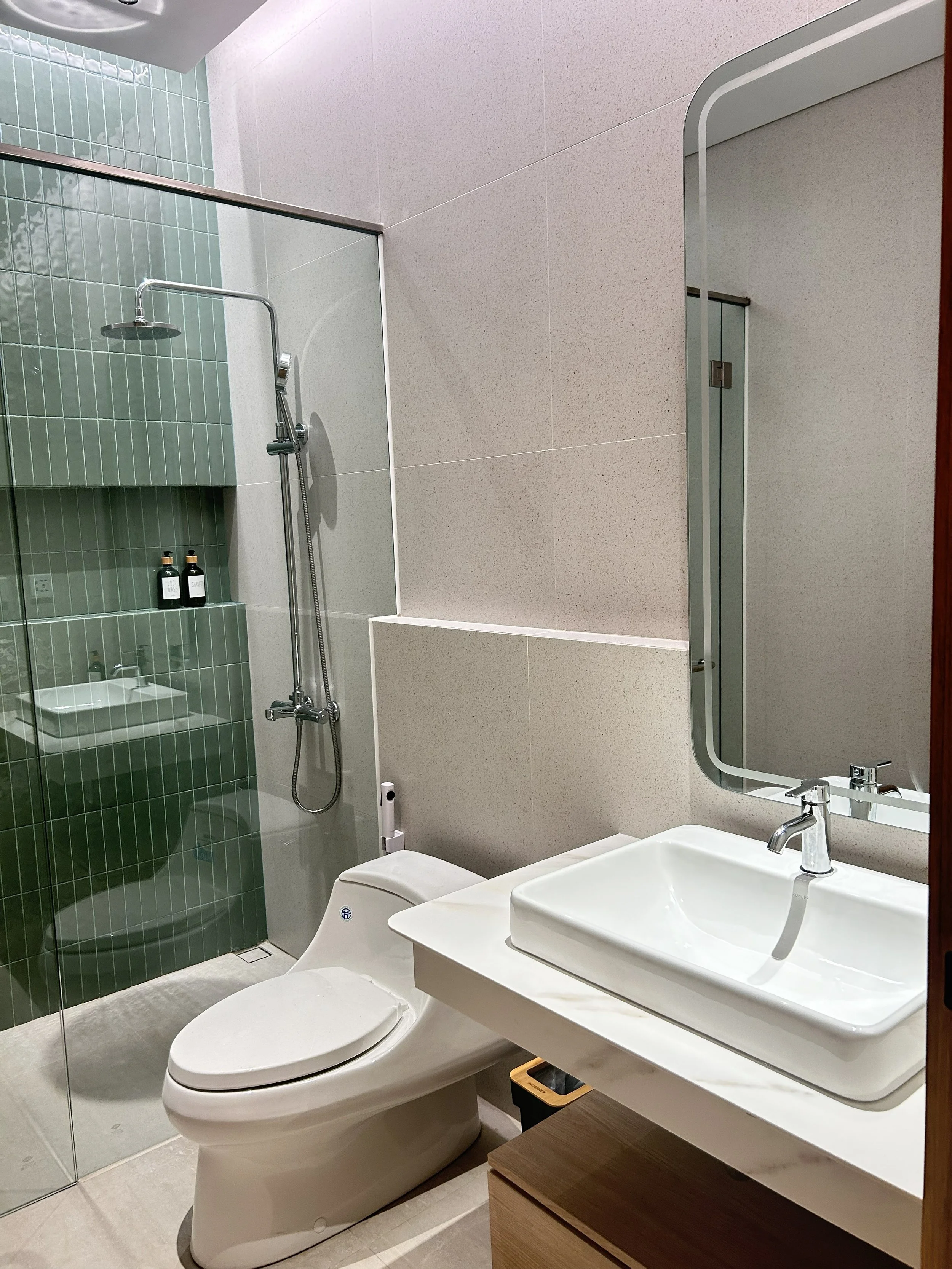 Modern bathroom with a glass shower enclosure featuring green tiles, a rainfall showerhead, and toiletries. Contains a white toilet and a white rectangular sink with a mirror above, set against beige walls and a wooden cabinet below the sink.
