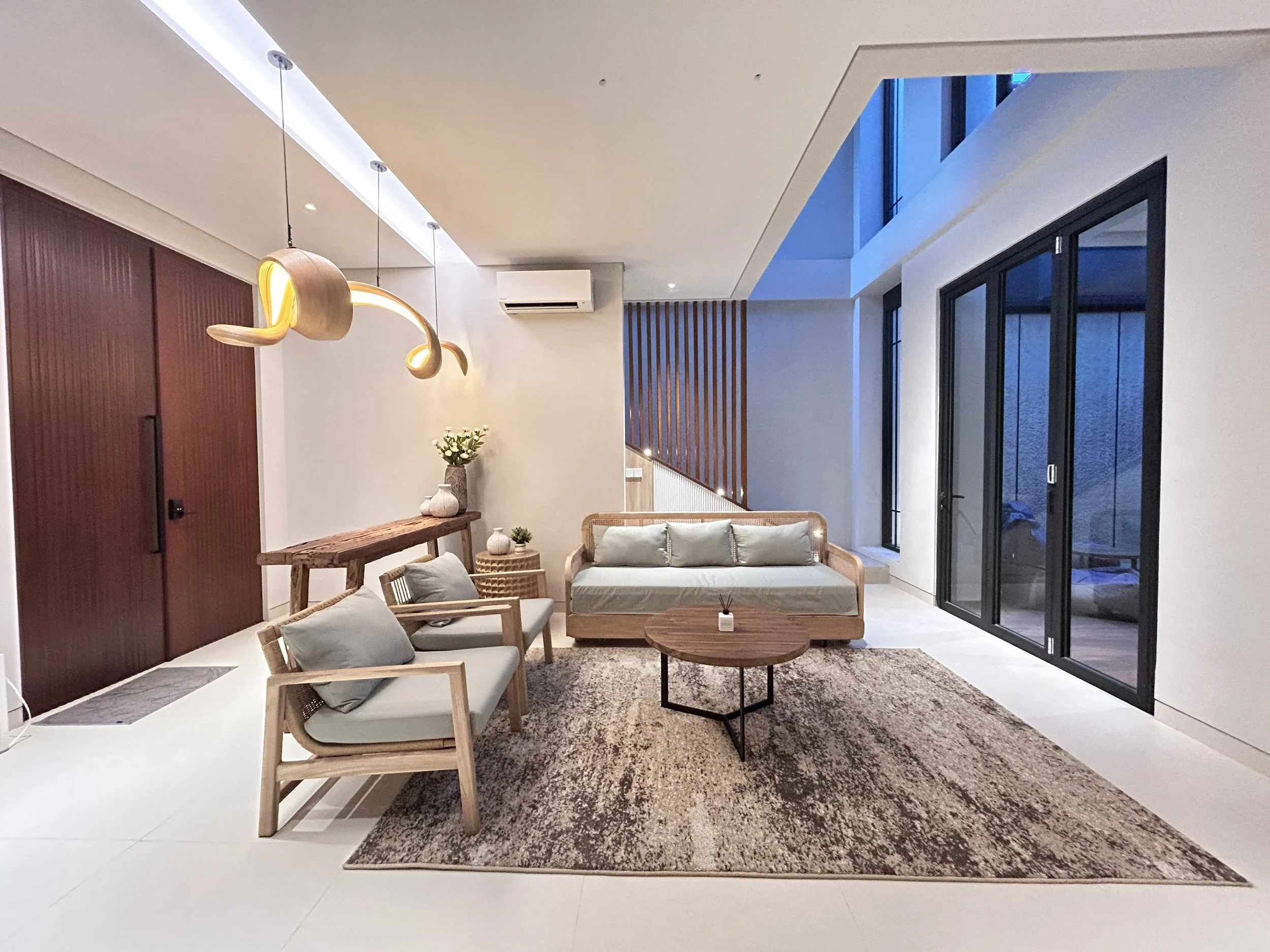 Modern living room with beige sofa, wooden chairs with gray cushions, a wooden coffee table, a textured beige and brown rug, large black-framed glass sliding doors, and contemporary pendant lighting.