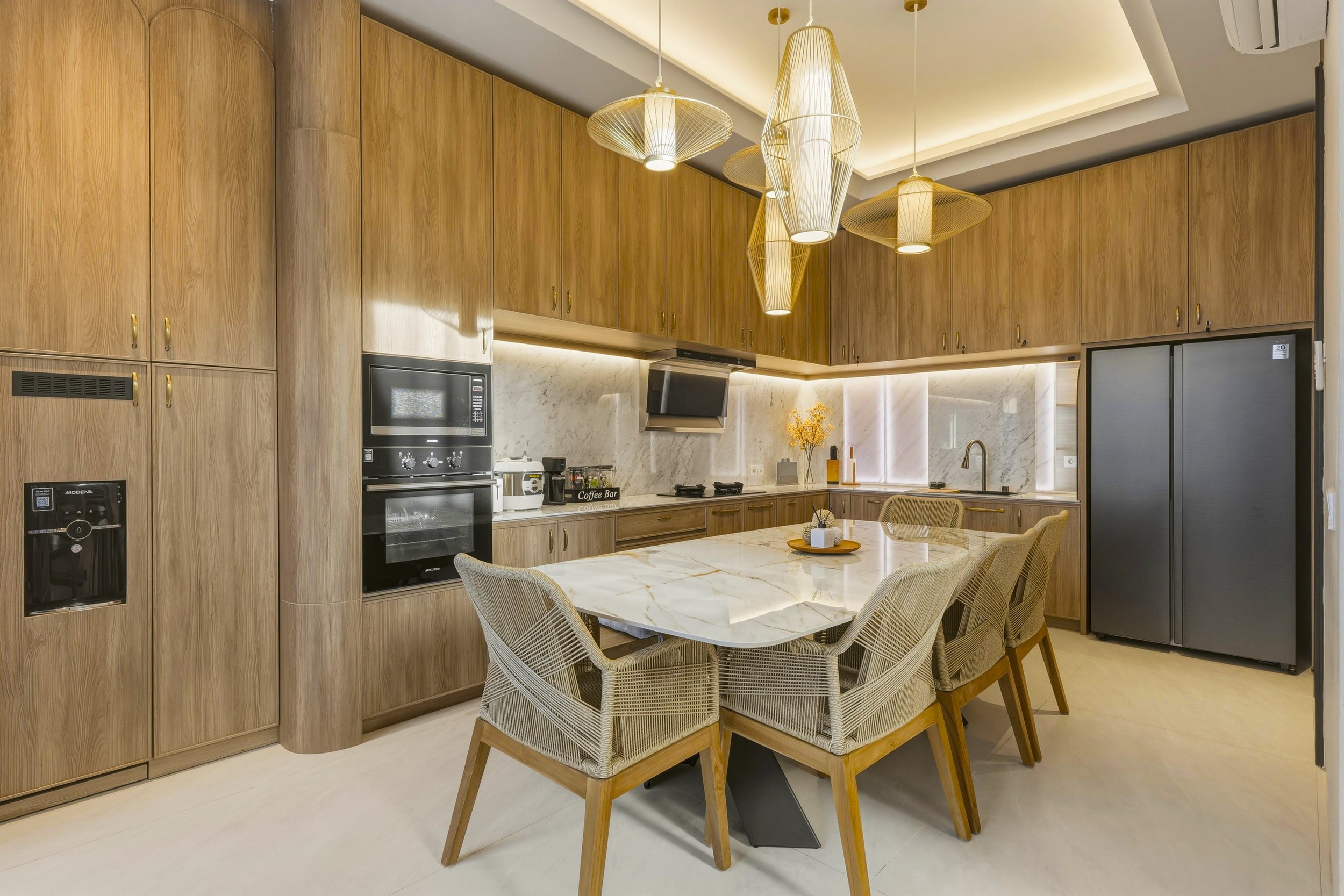 A modern kitchen with wooden cabinetry, a marble island table, and beige wicker chairs. Pendant lights hang from the ceiling, and there is a microwave, coffee machine, and refrigerator visible.