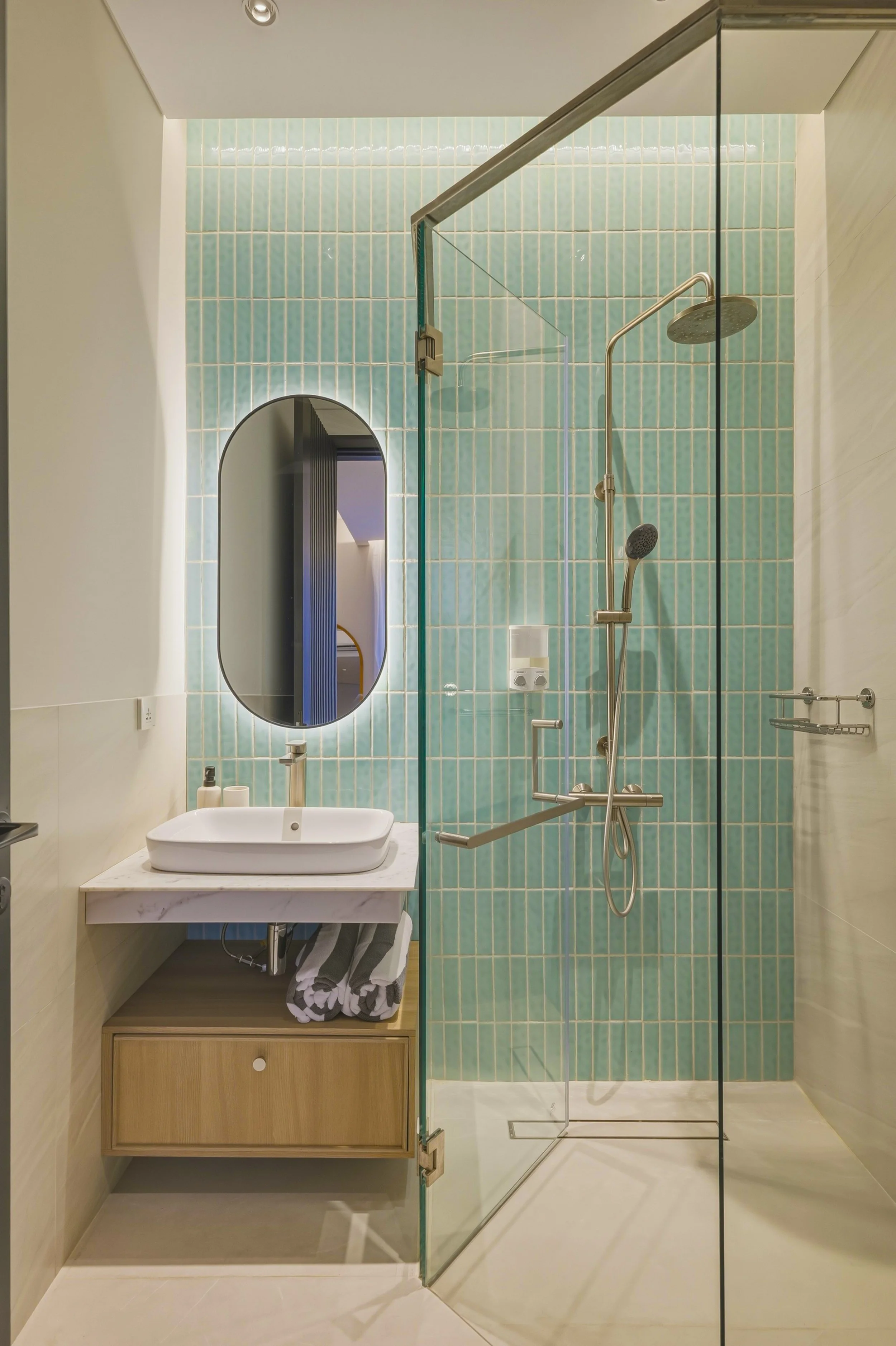 A modern bathroom with a walk-in shower with blue-green tiles, a glass door, a shower head on a vertical bar, and a white rounded sink on a marble countertop. There is a large oval mirror above the sink, a soap dispenser, and a wooden cabinet with a 