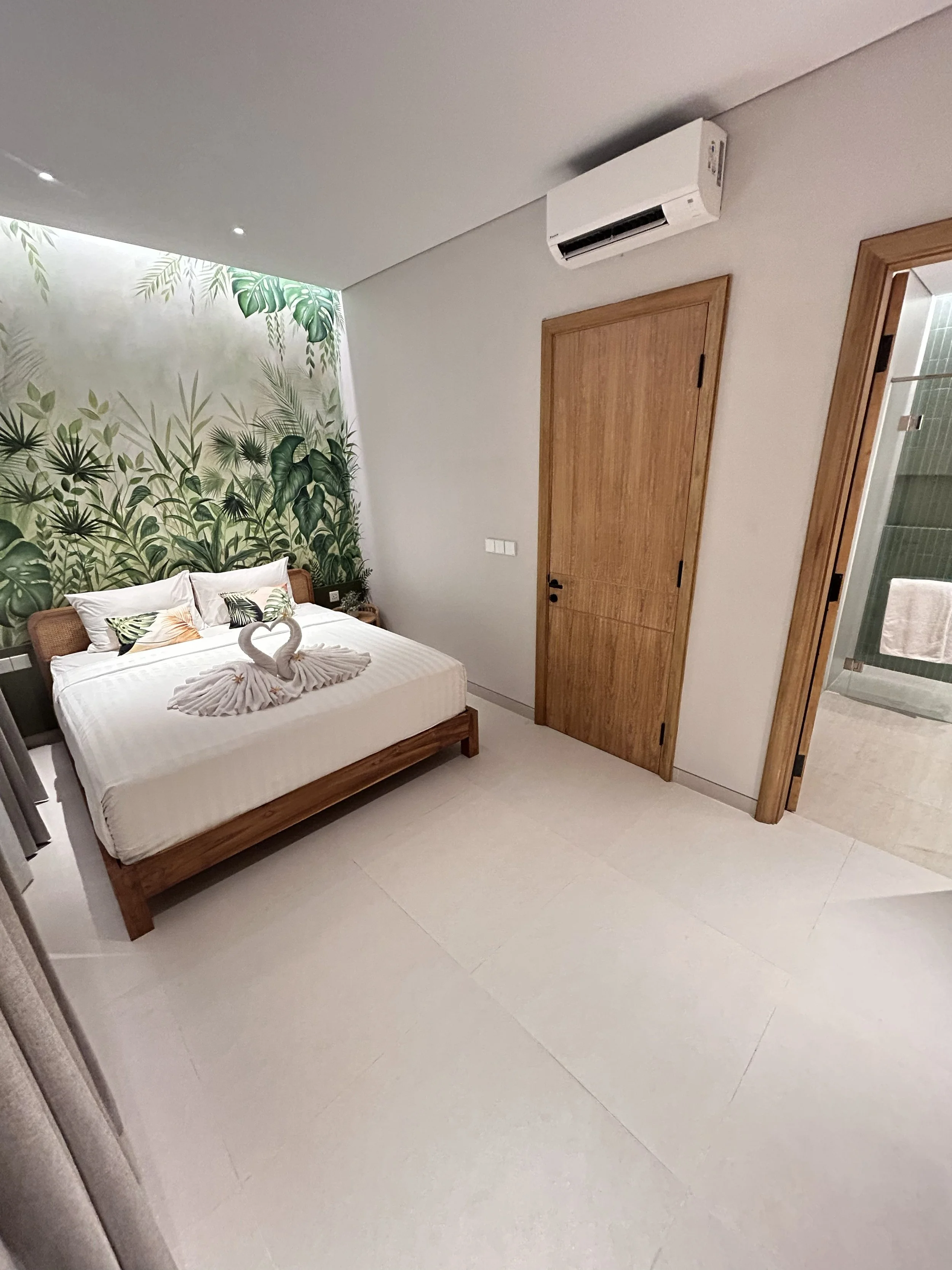 A bedroom with a bed, a tropical leaf wallpaper mural, an air conditioning unit, a wooden door, and an adjacent bathroom with green tiles visible through the doorway.