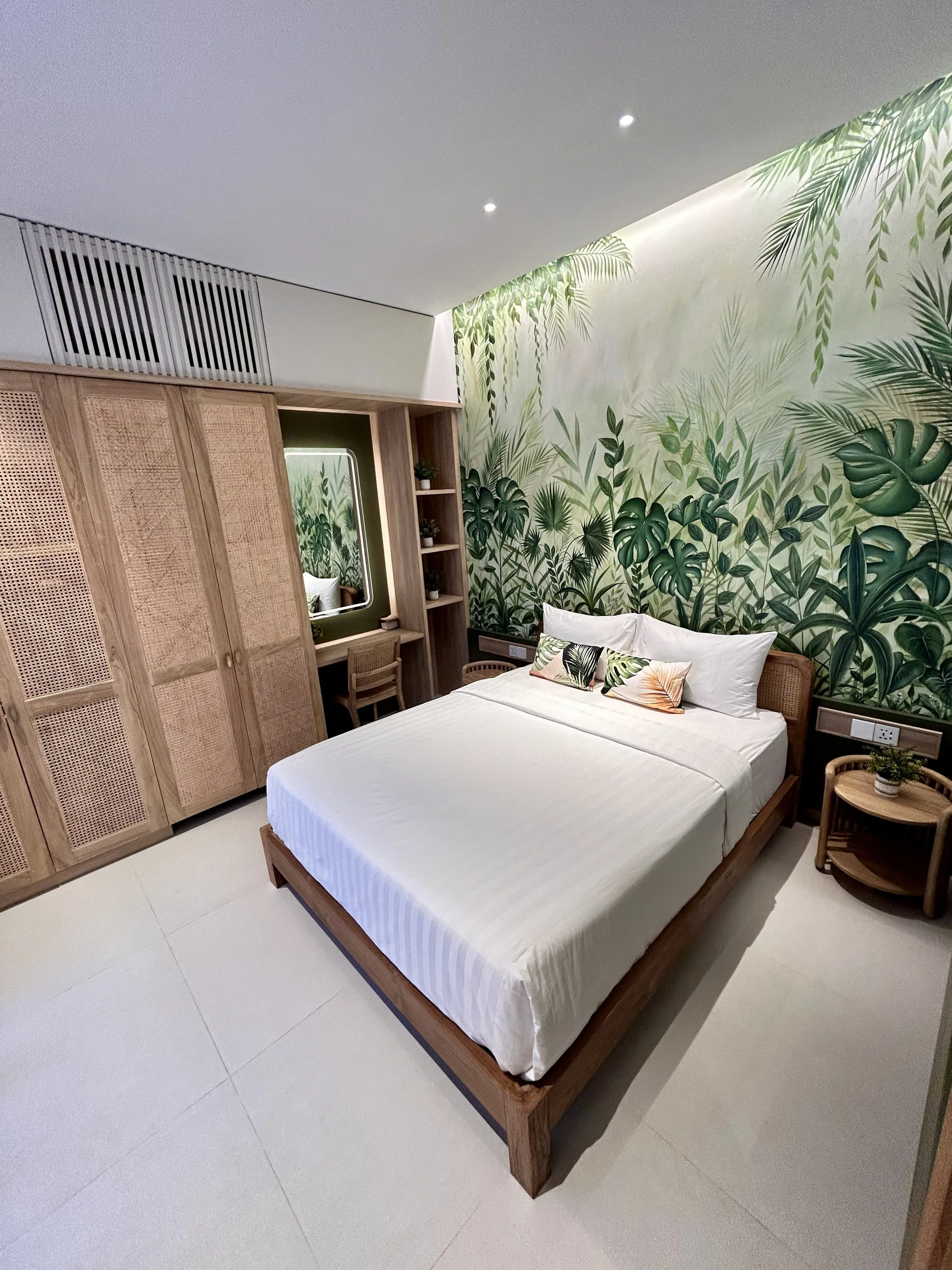 A hotel room with a wooden bed frame, white bedding, and tropical-themed pillows. There is a large green leafy mural on the wall behind the bed. To the right, a small wooden side table with a plant and a woven chair. On the left, a wooden wardrobe wi
