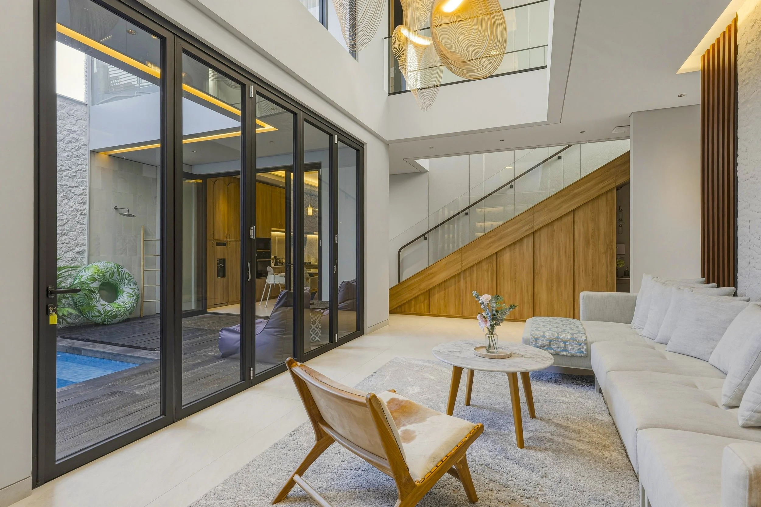 Modern living room with a beige sofa, a wooden chair, a marble coffee table with a vase of flowers, and sliding glass doors leading to an outdoor area with a pool and patio.