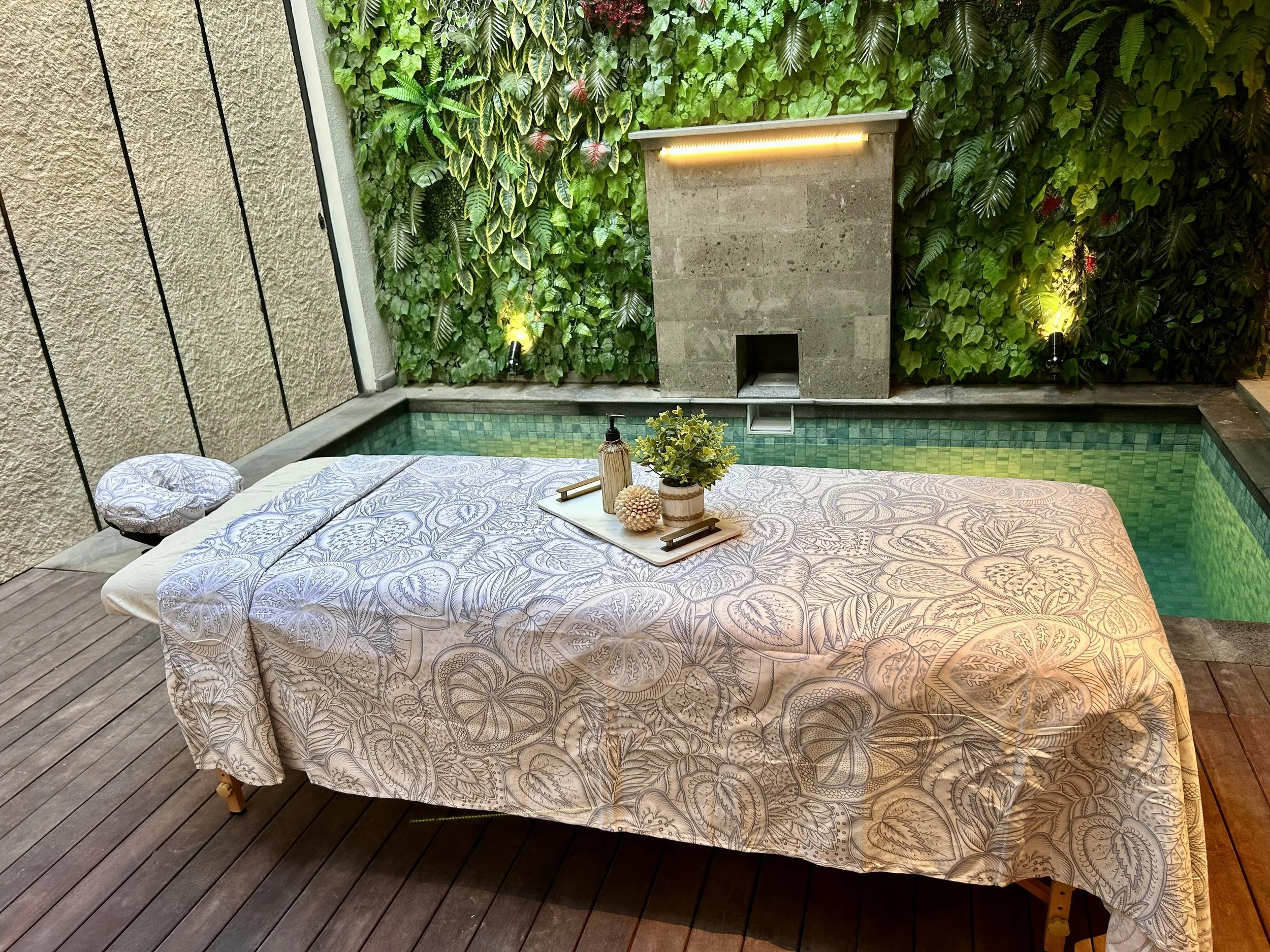Outdoor patio with a bed covered in a patterned beige and purple bedspread, a tray with decor and greenery, wooden deck, lush green wall with leafy plants, garden lights, wall and floor surfaces.