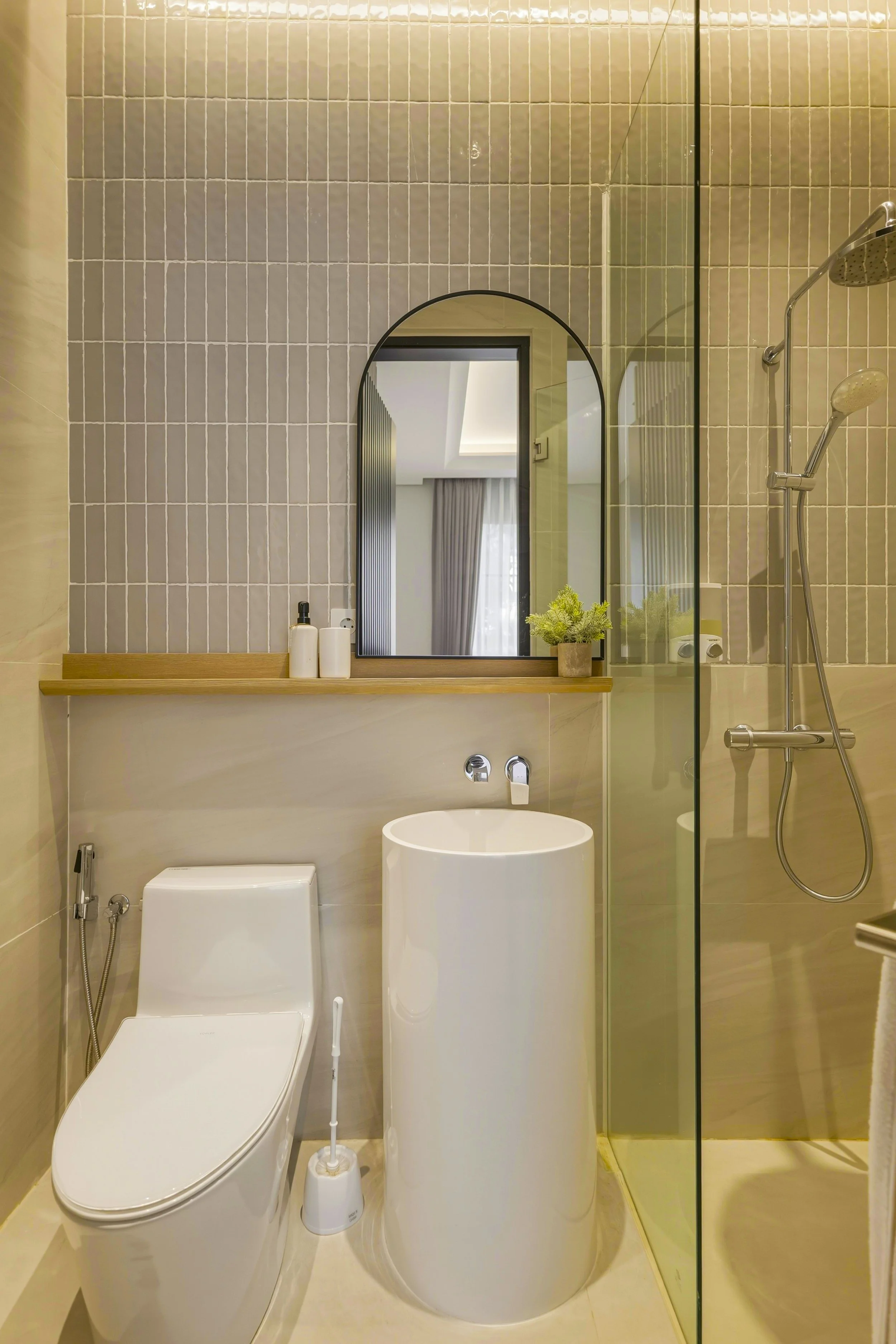 Modern bathroom featuring a white bidet, a toilet brush, a floating wooden shelf with bottles, a mirror, beige tiled walls, a small green plant, and a glass shower enclosure with dual shower heads.