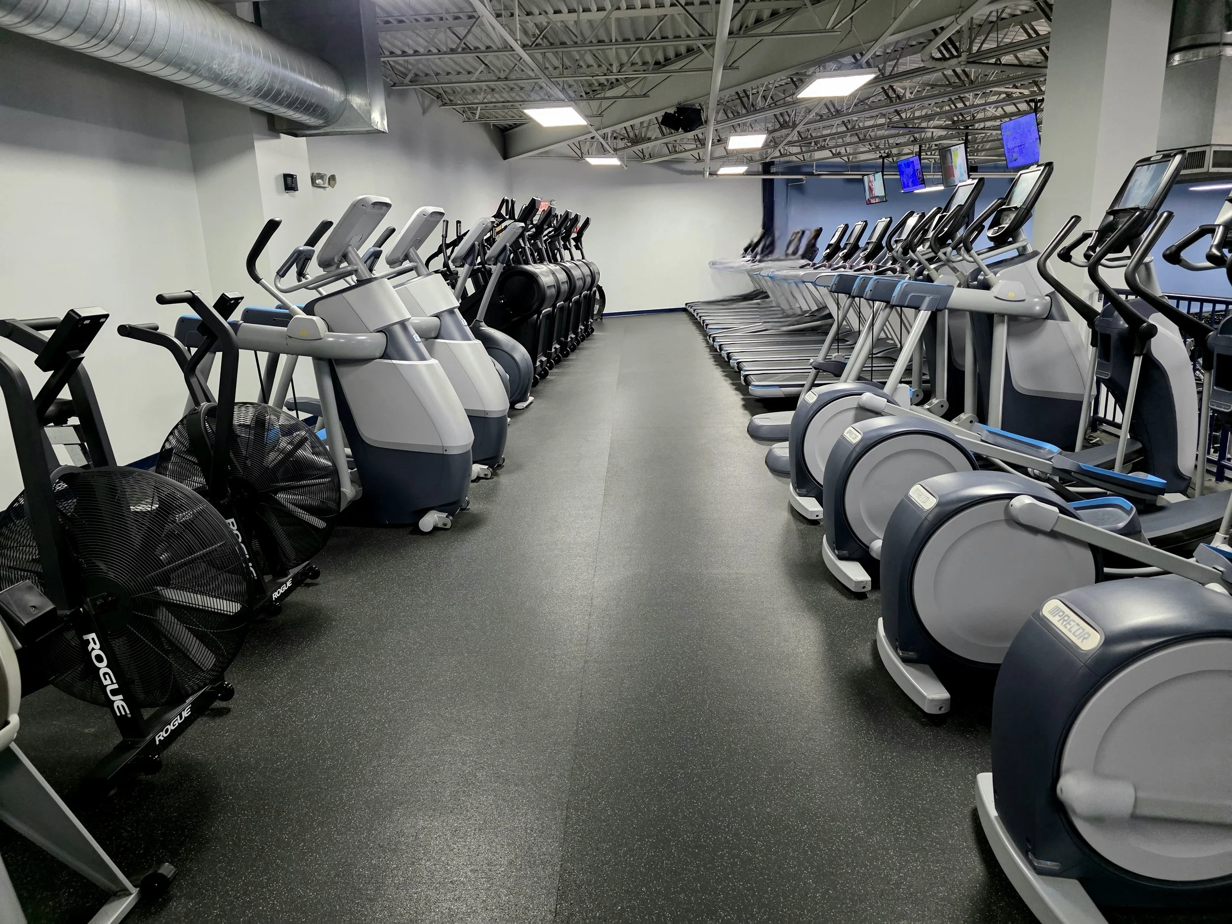 Empty gym with cardio equipment including stationary bikes, elliptical trainers, and treadmills lined up in rows.