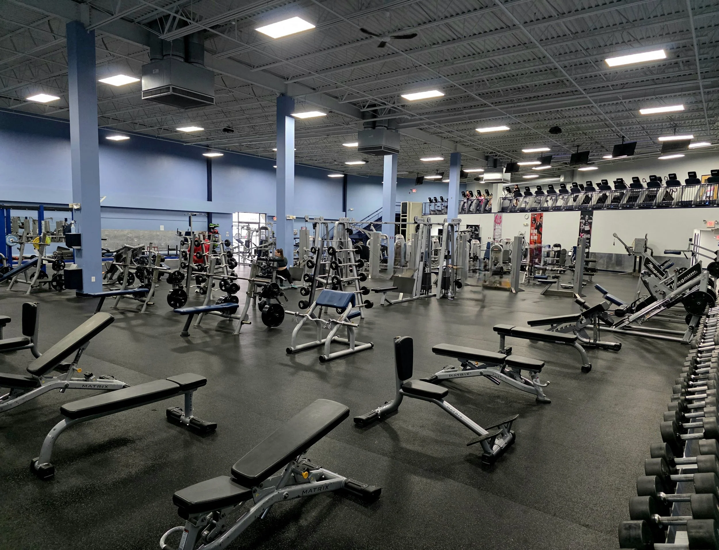 Interior of a large gym with various weightlifting equipment, benches, and a cardio area with treadmills on an upper level.