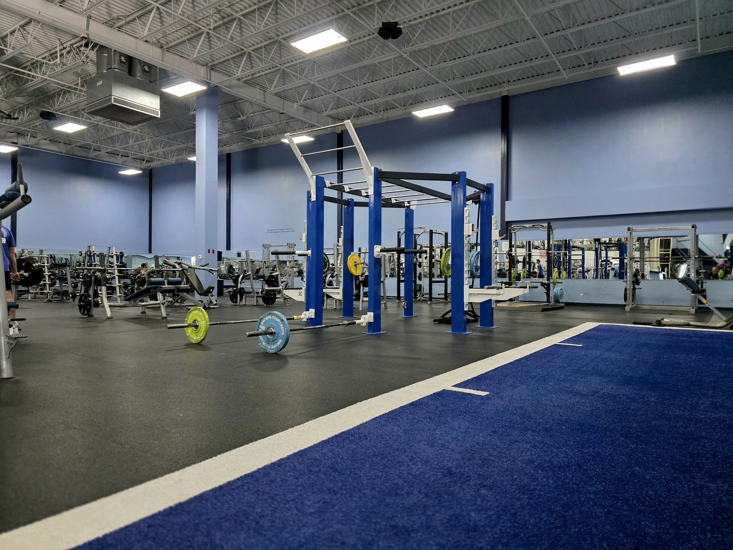 Interior of a gym with various weightlifting equipment, including barbells, weight plates, and squat racks, with blue walls and a large mirror along one side.